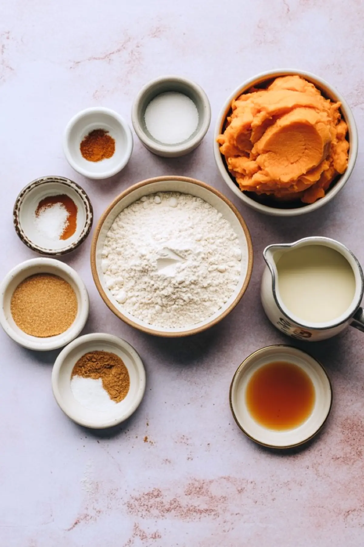 Flat lay of sweet potato pancake ingredients including mashed sweet potatoes, flour, spices, sugar, baking powder, milk, and vanilla extract in ceramic bowls.
