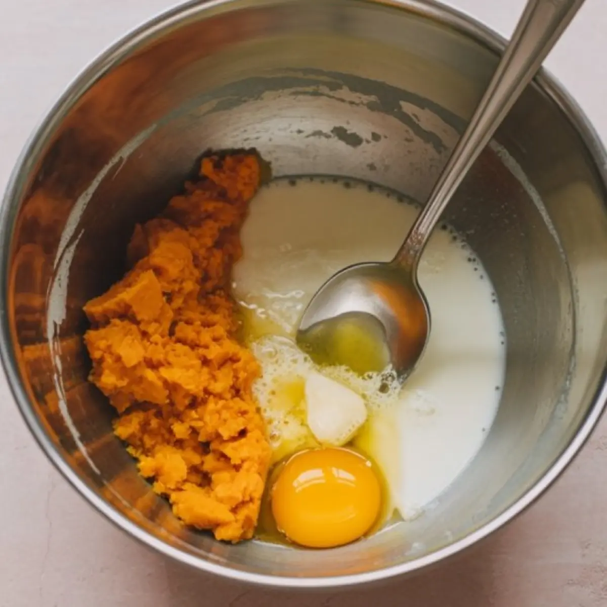 Stainless steel bowl containing mashed sweet potato, raw egg, melted butter, and milk being prepared for sweet potato pancake batter with a spoon.