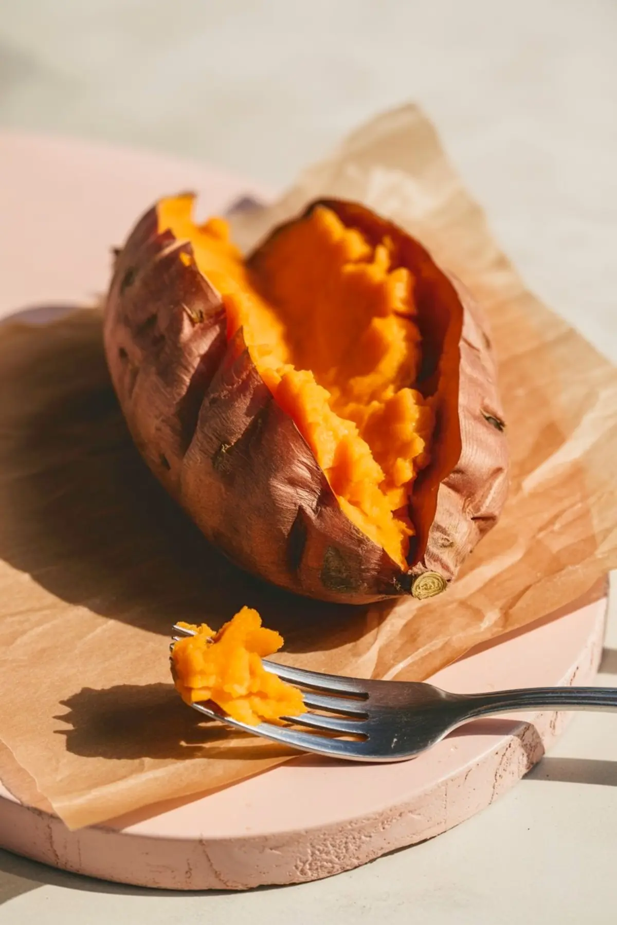 Baked sweet potato split open with a forkful of orange flesh on parchment paper under natural light.