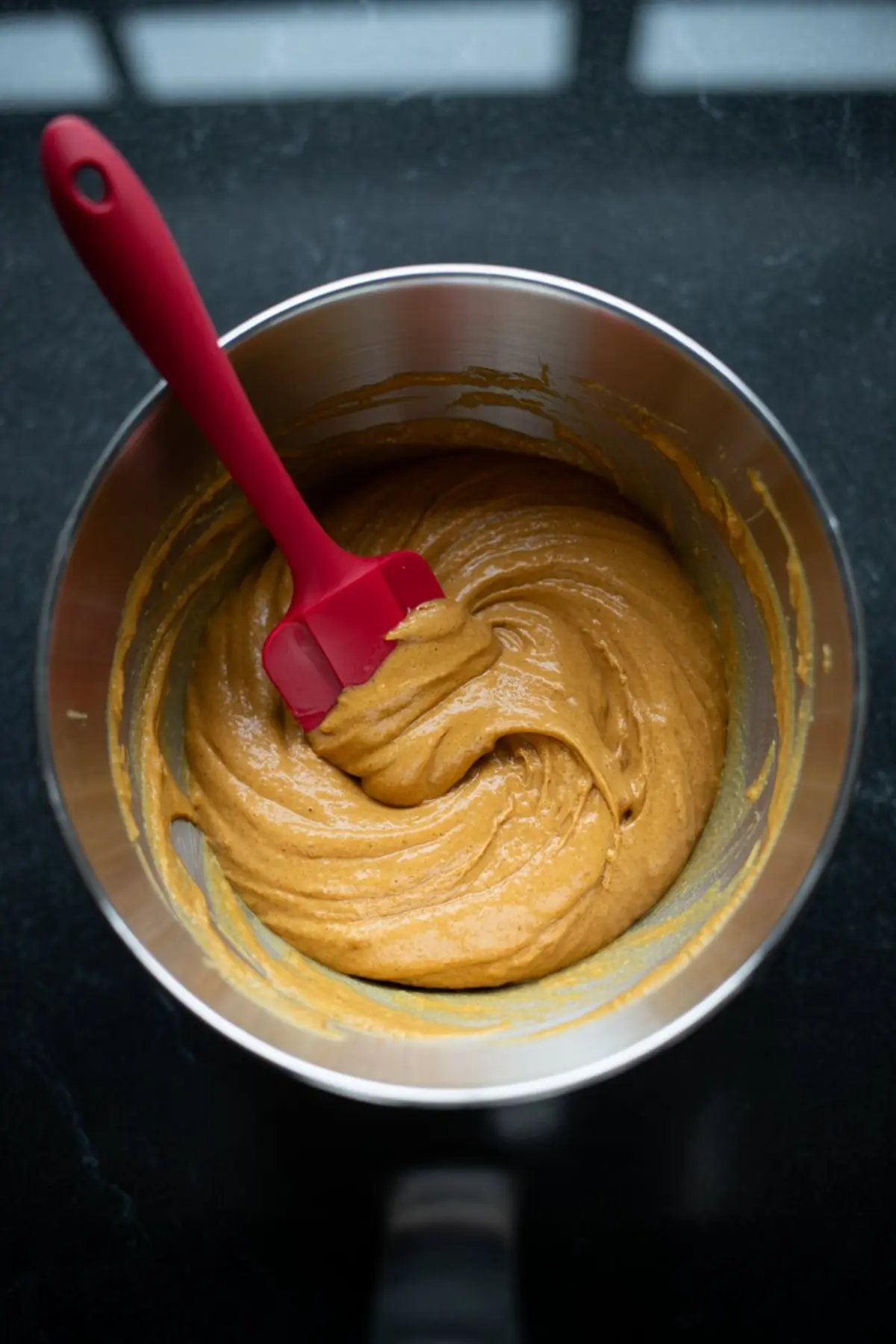Thick vegan pumpkin bread batter being mixed in a metal bowl with a red silicone spatula, showing a smooth and creamy texture ready for baking.