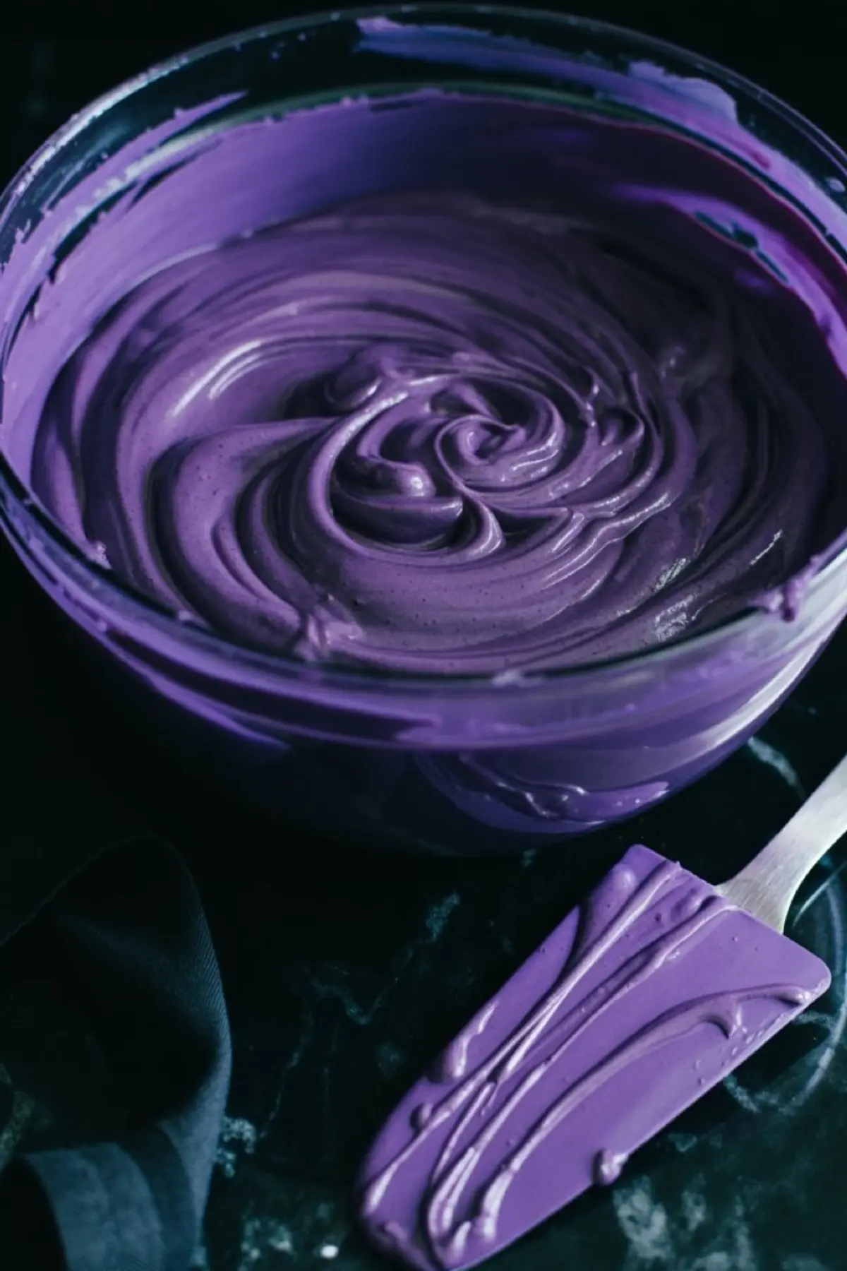 A large bowl of whipped purple cheesecake batter with a matching spatula coated in the same colorful mixture, placed on a dark marble counter.