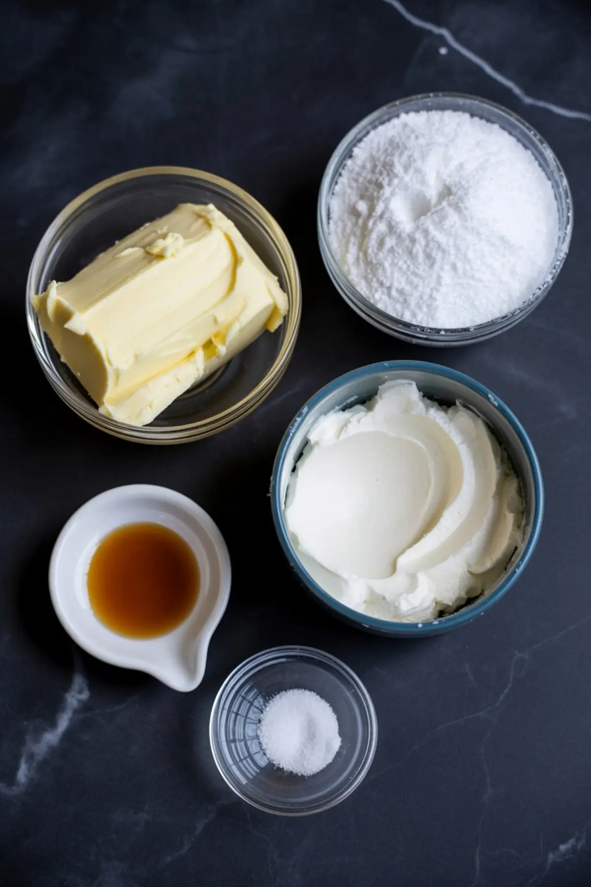 Glass bowls on a black surface filled with ingredients for buttercream frosting including unsalted butter, powdered sugar, vanilla extract, salt, and heavy cream.
