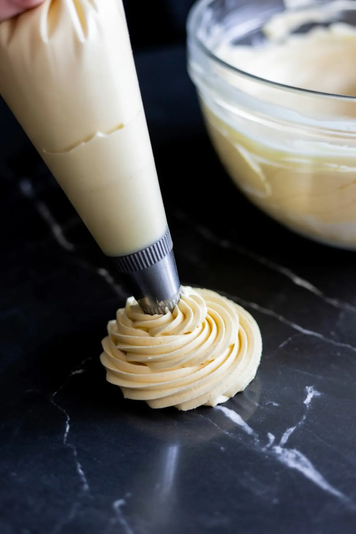 Piping bag with a star tip forming a swirl of smooth vanilla buttercream frosting on a dark marble countertop.
