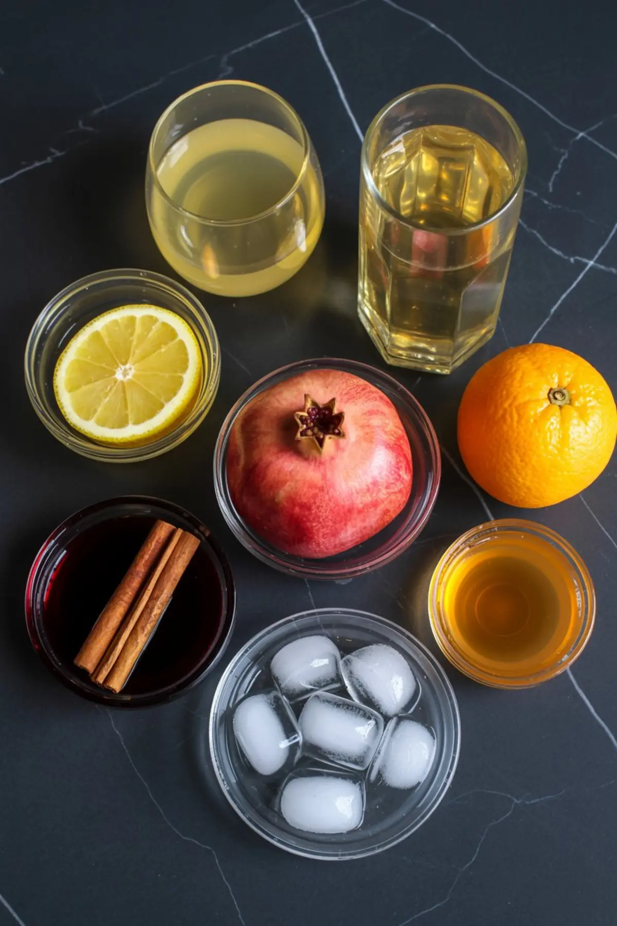 Overhead view of assorted apple cider sangria ingredients on a dark marble surface, including fresh lemon slices, whole orange, pomegranate, cinnamon sticks, ice cubes, and various juices in glass bowls and glasses.
