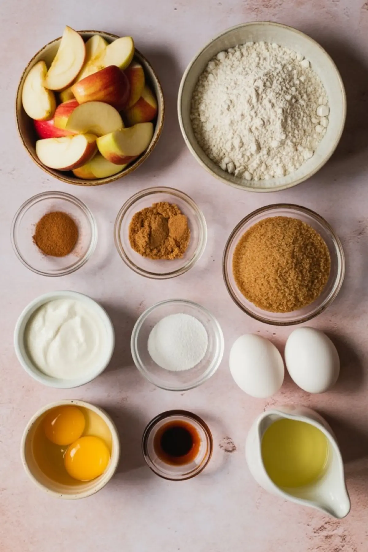 Flat lay of apple cinnamon muffin ingredients including sliced apples, all-purpose flour, brown sugar, granulated sugar, ground cinnamon, nutmeg, eggs, vanilla extract, yogurt, and oil arranged on a light pink surface.