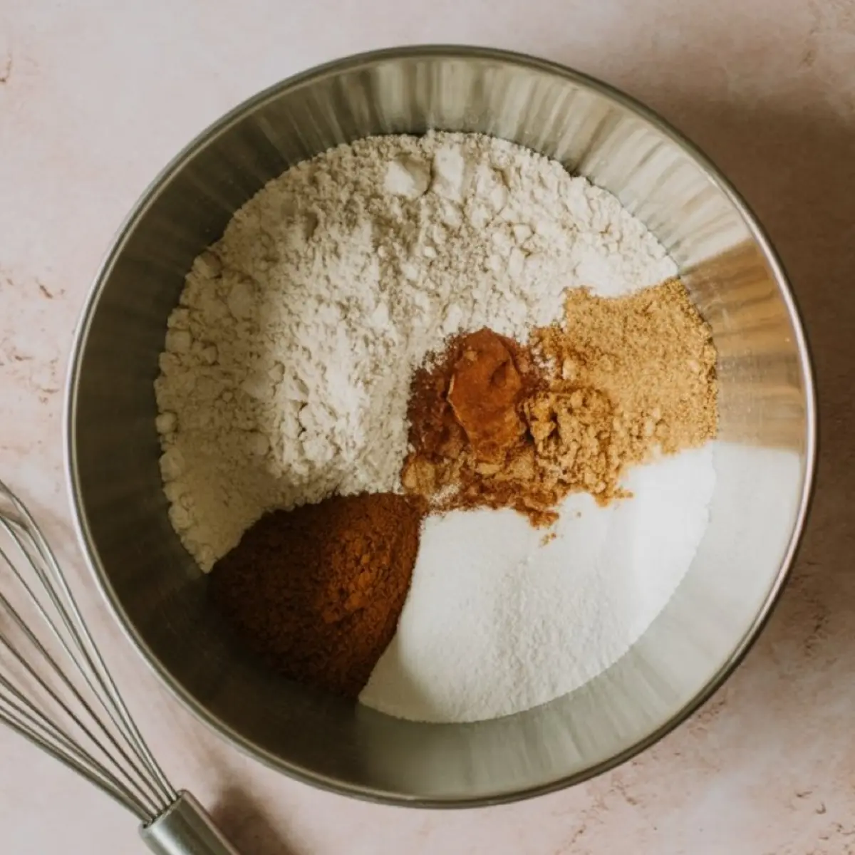 Mixing bowl with dry muffin ingredients including flour, granulated sugar, brown sugar, cinnamon, and nutmeg, placed beside a metal whisk on a pink surface.