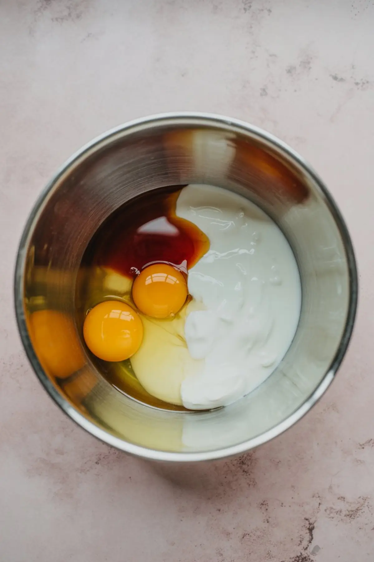 Overhead view of a metal mixing bowl containing eggs, yogurt, vanilla extract, and oil, ready to be mixed for apple cinnamon muffin batter.