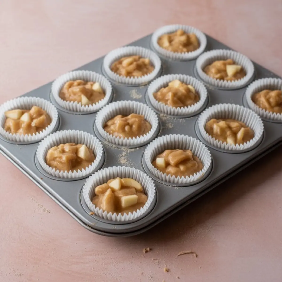 Muffin tin filled with apple muffin batter topped with chopped apple pieces, placed in white paper liners, ready to be baked.