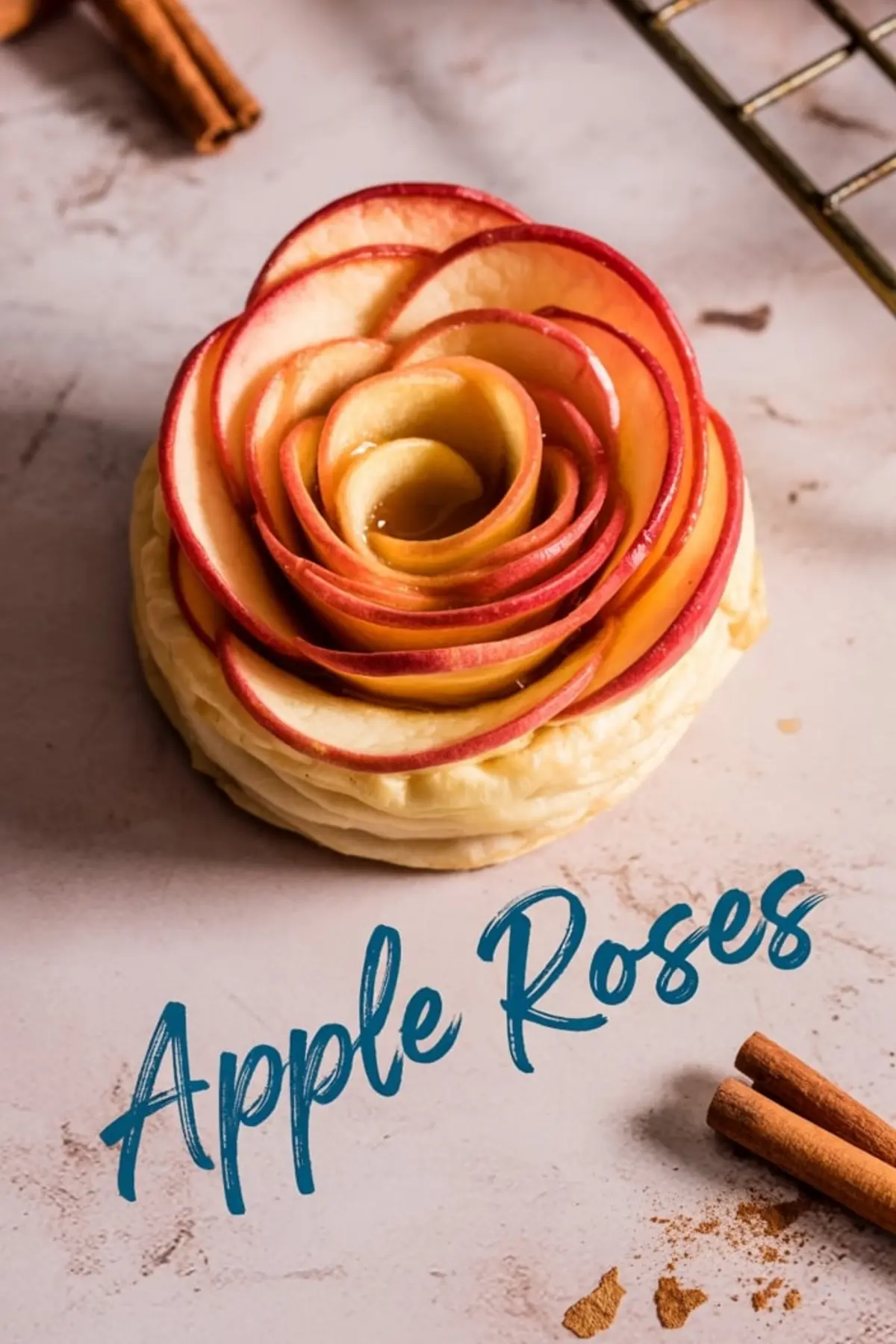 Overhead shot of an apple rose pastry with delicately layered red apple slices on a puff pastry base, styled with cinnamon sticks and the text “Apple Roses” in blue cursive font on a marble-textured surface.