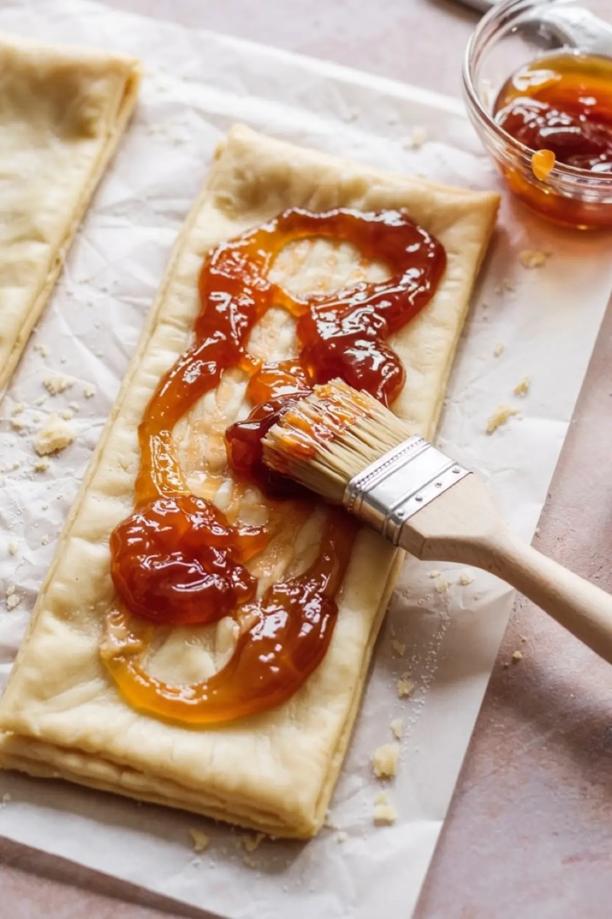 Close-up of apricot jam being brushed onto a rectangular sheet of puff pastry with a pastry brush, showing the glossy jam texture and layered dough on parchment paper.