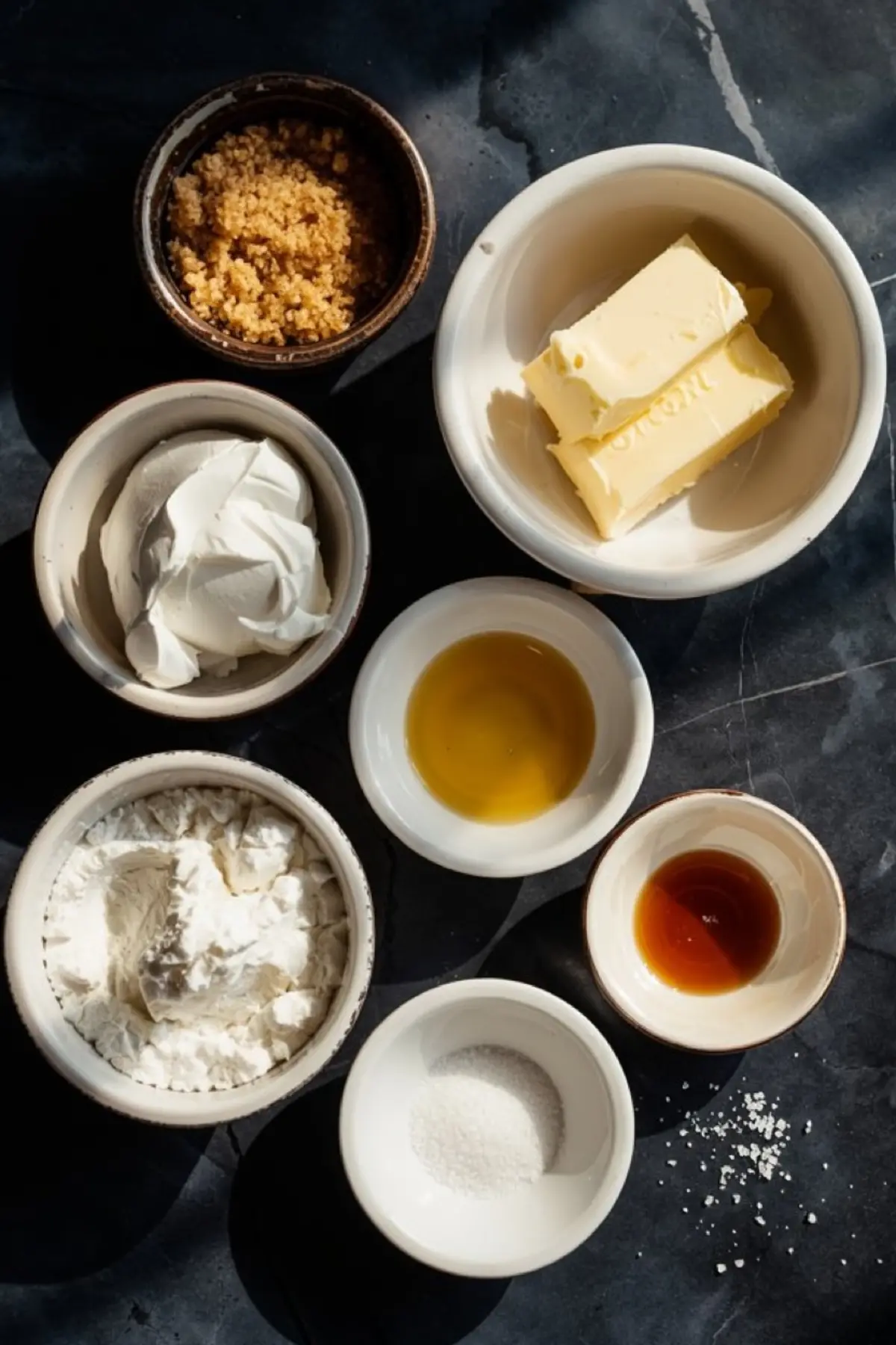 Overhead view of individual ingredients for brown sugar frosting arranged in small bowls, including butter, sour cream, brown sugar, powdered sugar, vanilla extract, and salt.
