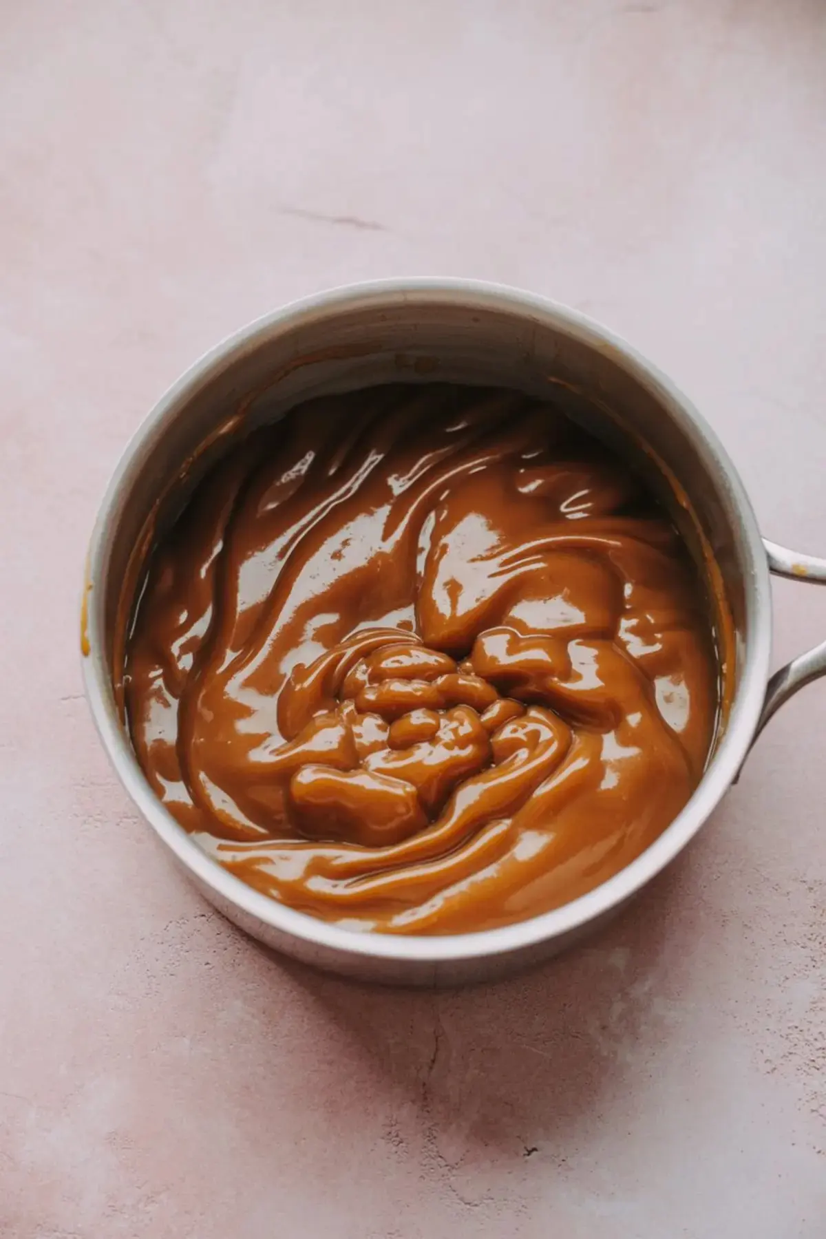 Thick homemade caramel sauce swirled in a saucepan on a light pink background, showing a glossy texture perfect for dessert toppings and snacks.
