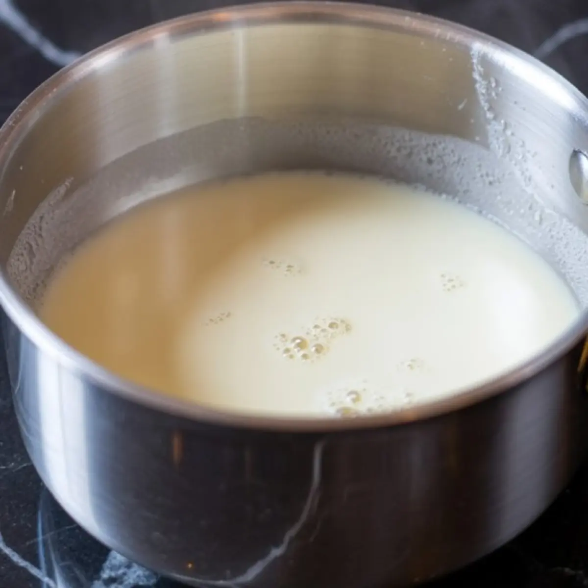 Heated heavy cream in a stainless steel saucepan with light bubbling on the surface, placed on a dark marble stove area.