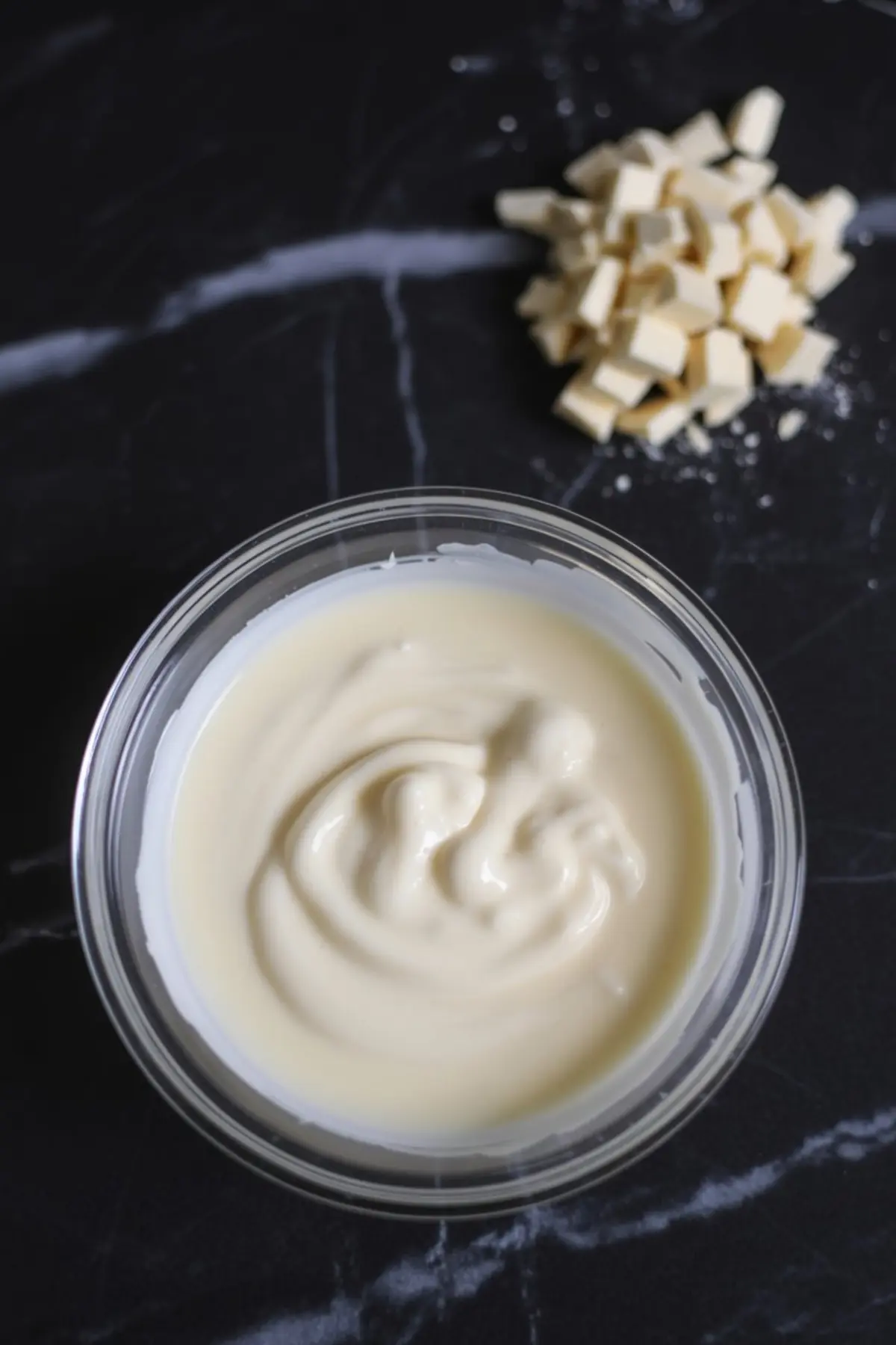 Bowl of smooth, melted white chocolate with chopped white chocolate pieces in the background, used for layered chocolate bark or Christmas dessert decorating.