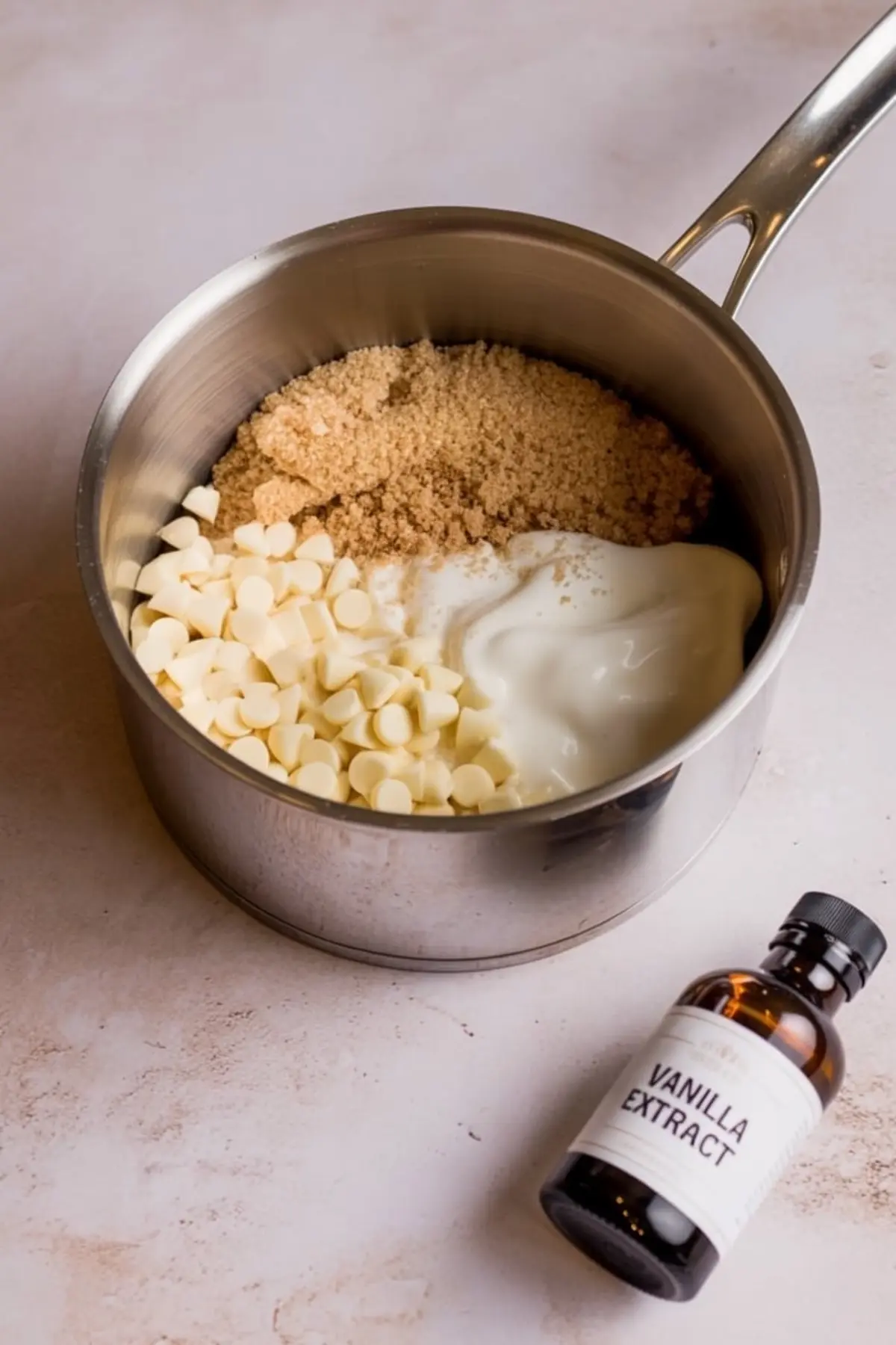 Saucepan filled with white chocolate chips, brown sugar, and thick cream next to a bottle of vanilla extract, showing ingredients for making caramel Christmas fudge.