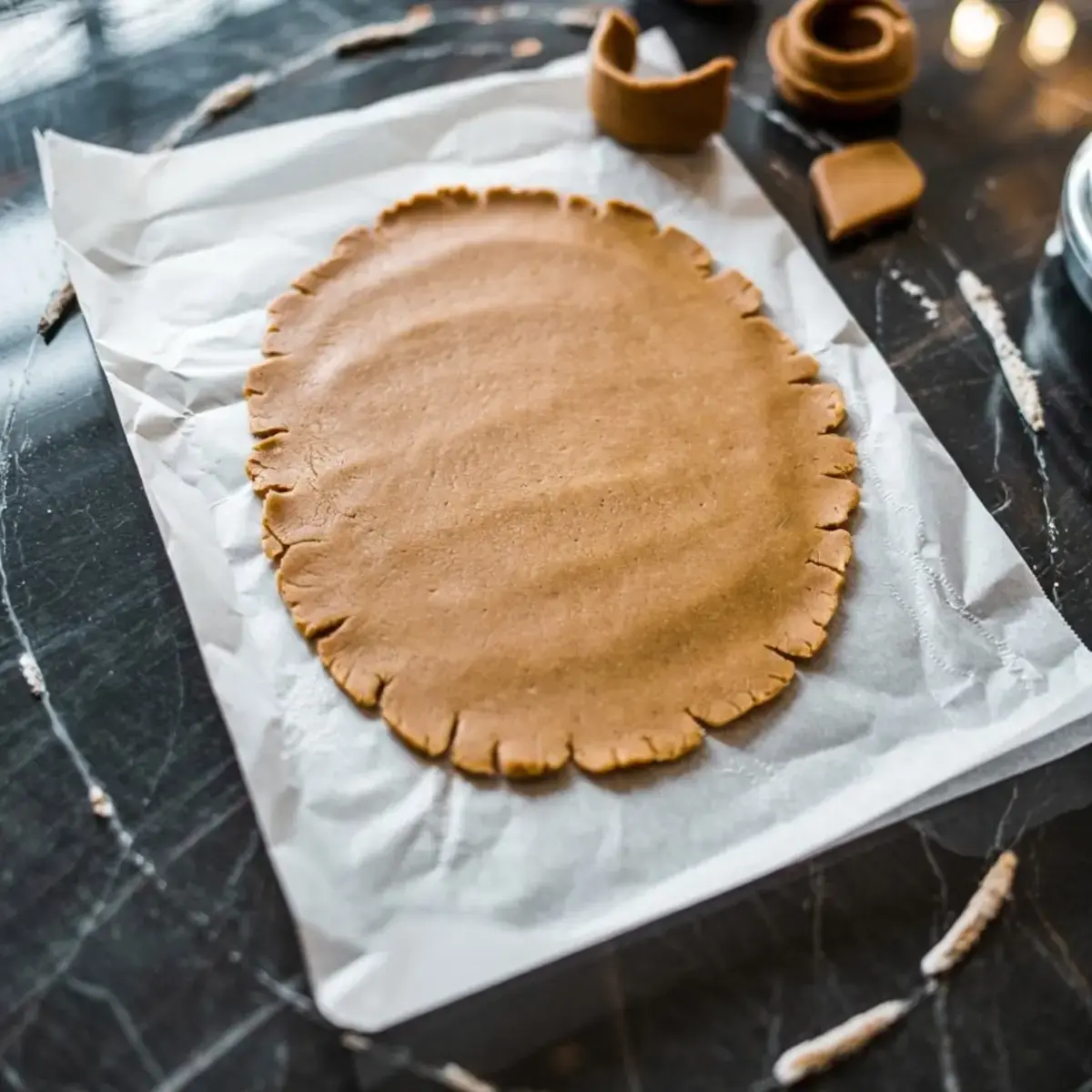 Rolled-out gingerbread dough on parchment paper, shaped into an oval with fluted edges, ready to be cut into cookie pieces for holiday baking.
