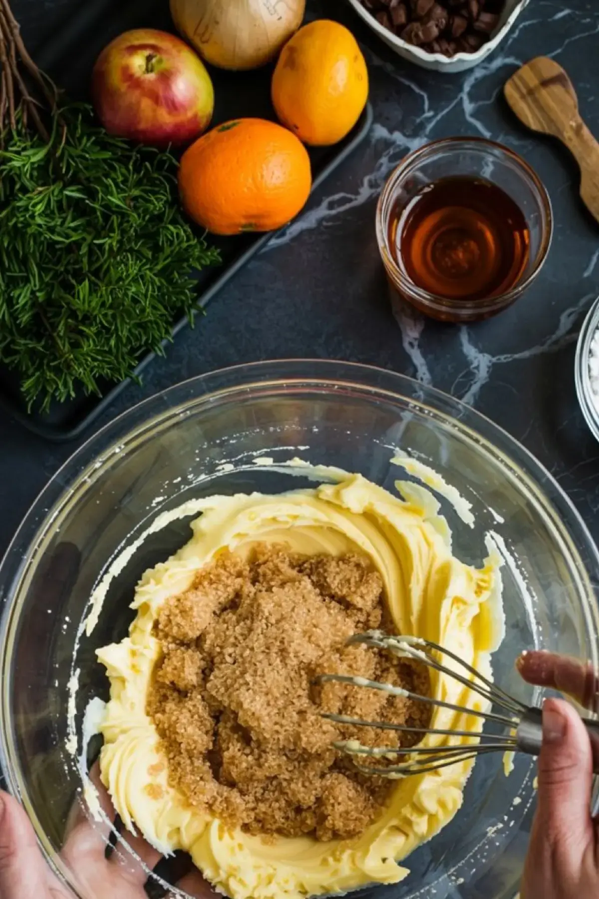 Butter and brown sugar being mixed in a glass bowl by hand with a whisk, surrounded by fresh oranges, apples, rosemary, and baking ingredients on a black kitchen counter.
