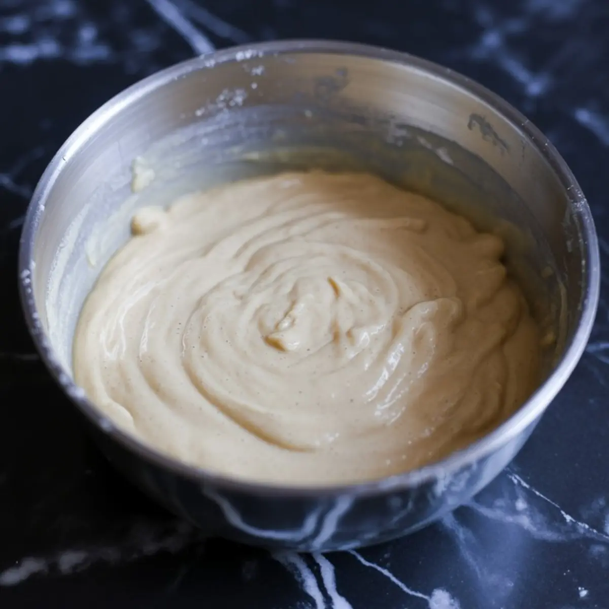 Smooth and creamy pancake batter swirled in a metal mixing bowl on a black marbled surface, ready to be cooked into fluffy pancakes.