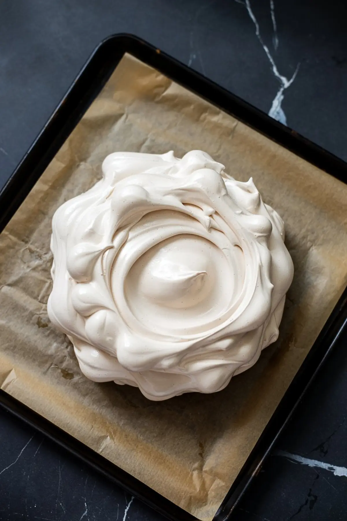 Unbaked pavlova meringue shaped into a circular nest with swirled peaks, resting on parchment paper in a baking tray, ready to be baked into a crisp and airy dessert base.

