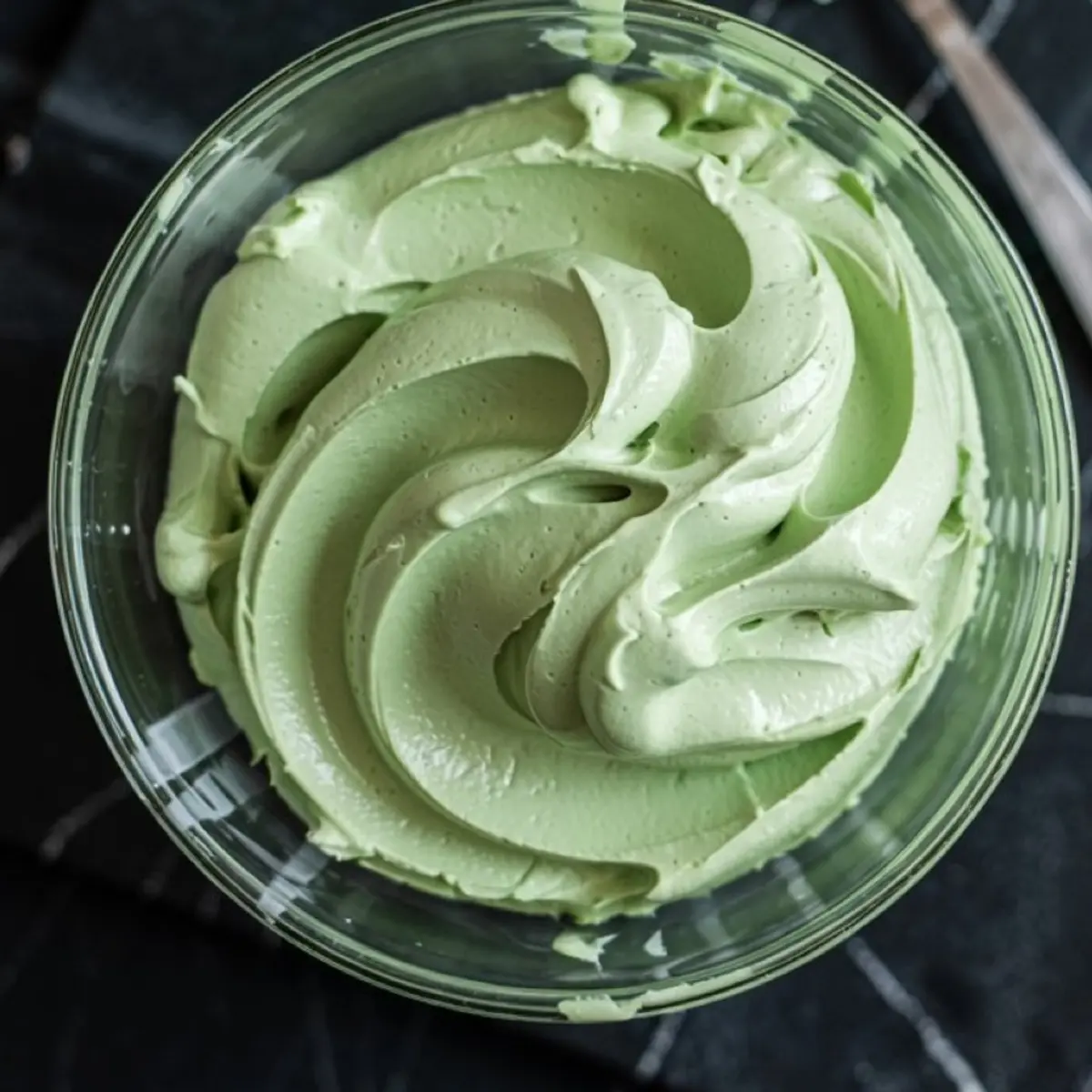 Thick swirl of pale green frosting in a glass bowl, whipped to a creamy consistency, on a black countertop.