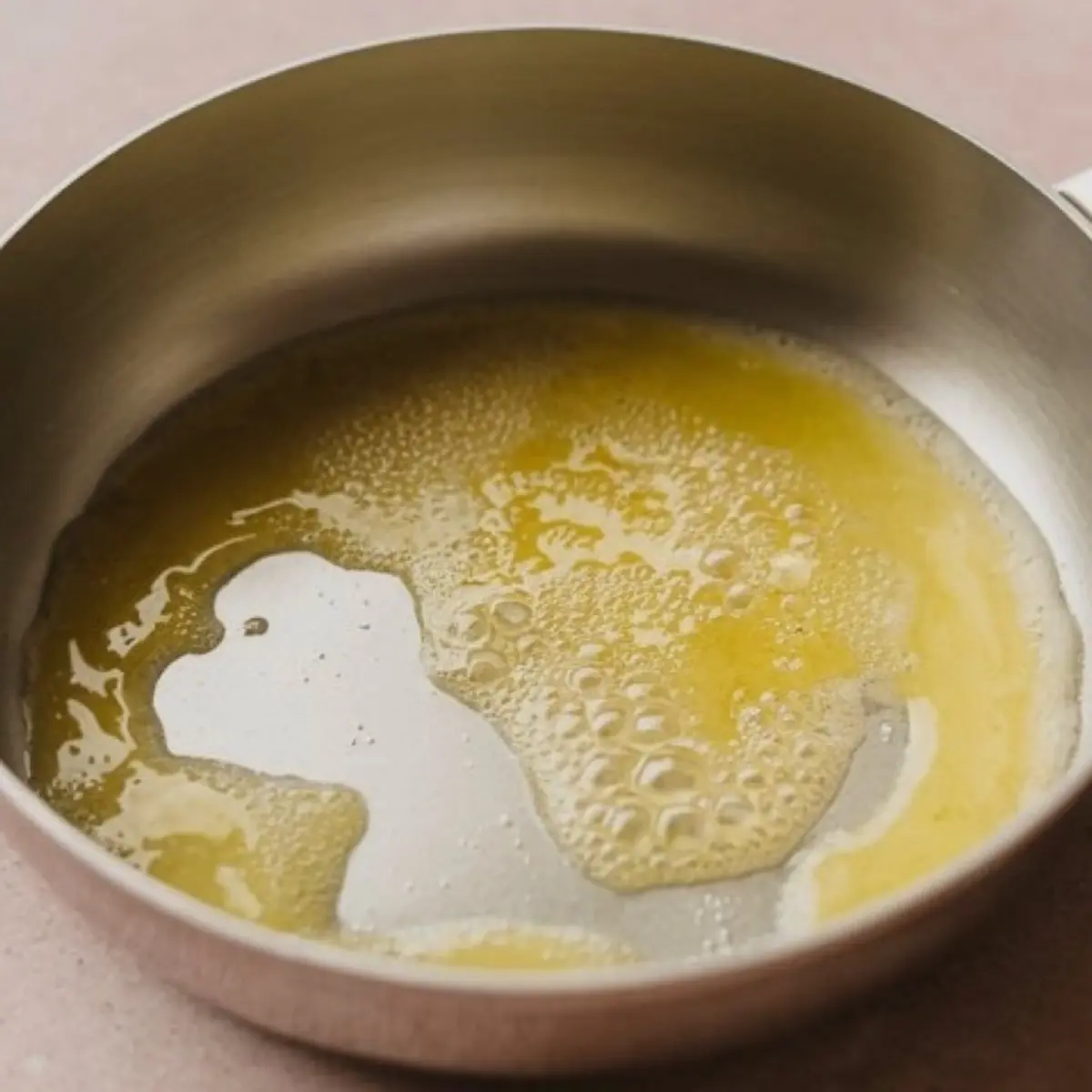 A close-up view of melted butter bubbling in a stainless steel pan, captured on a light countertop with a soft pink hue.
