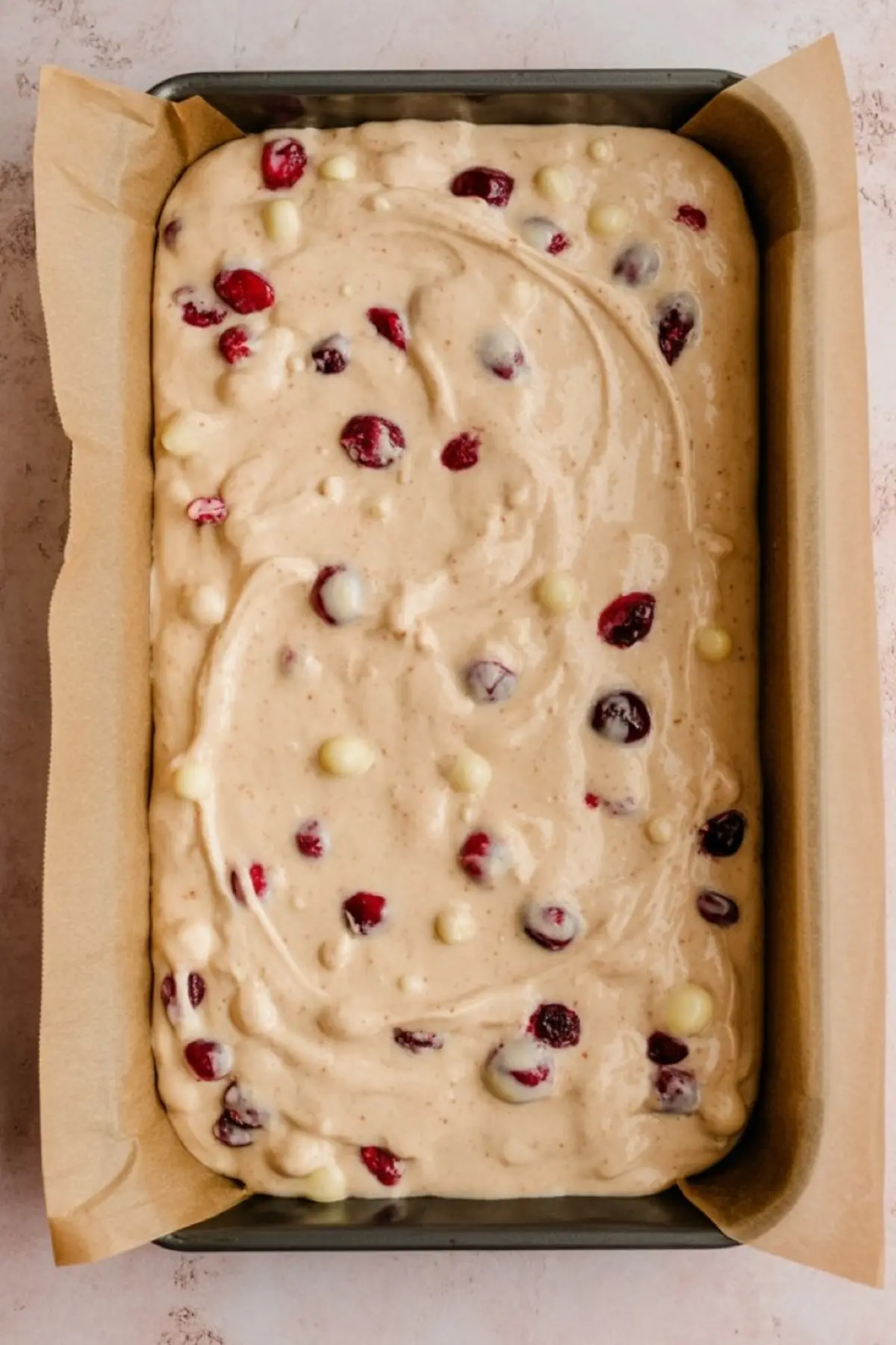 Unbaked cranberry blondie batter in a parchment-lined pan, dotted with whole cranberries and white chocolate chips, ready for the oven. The creamy mixture swirls with festive red and white colors.
