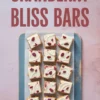 Top view of sliced cranberry bliss bars arranged on a parchment-lined tray, topped with smooth white chocolate frosting and dried cranberries. Text overlay highlights the dessert name for easy Pinterest recognition.