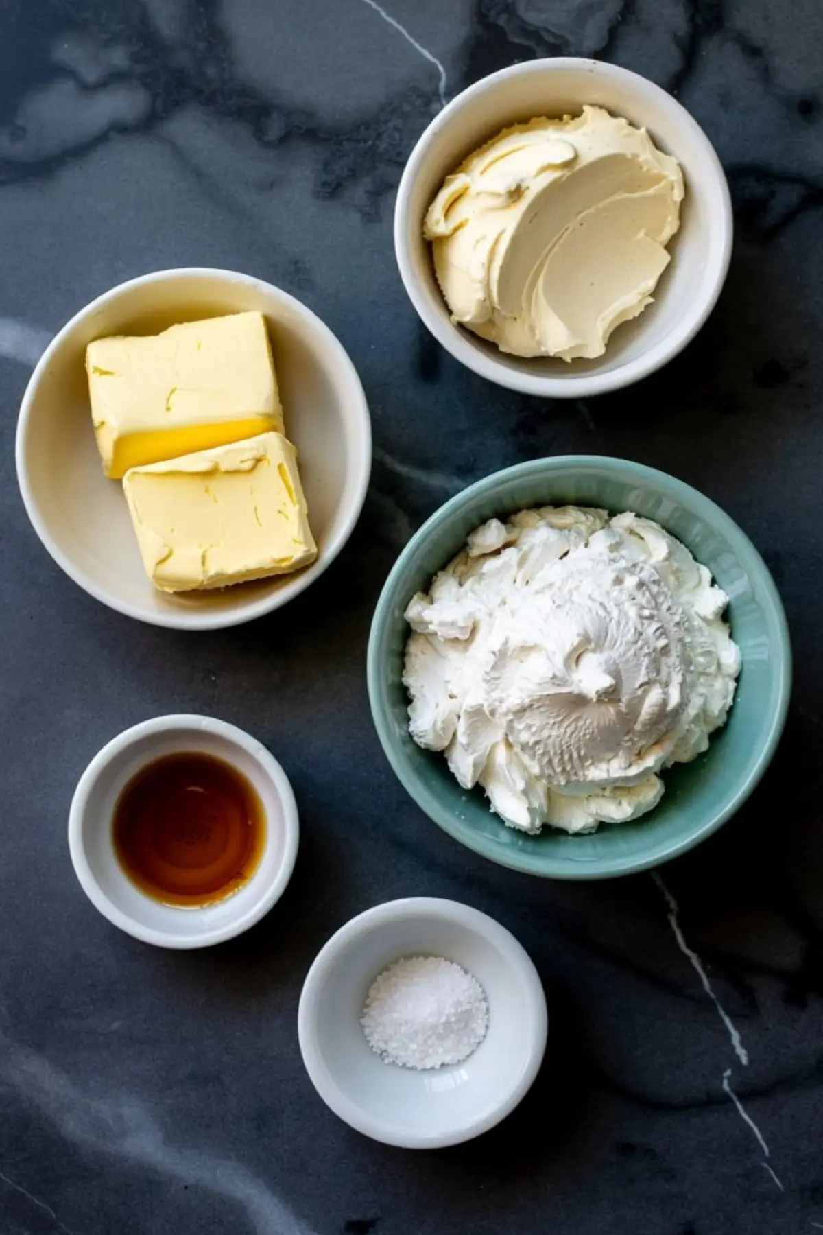 Overhead view of small bowls filled with cream cheese, butter, whipped cream, vanilla extract, and salt, arranged on a black marble countertop for a baking recipe.