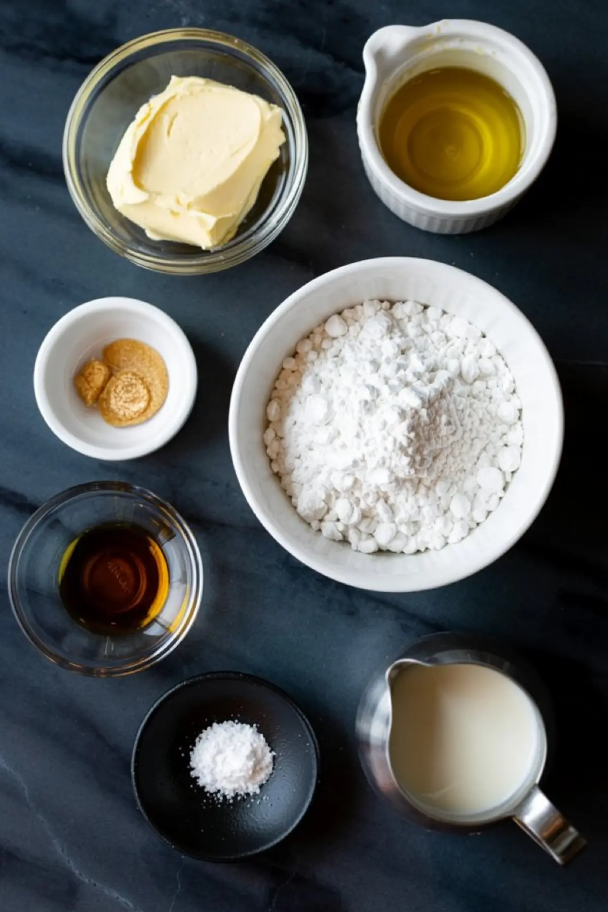 Flat lay of dairy-free frosting ingredients on a dark countertop, including vegan butter, olive oil, powdered sugar, plant-based milk, maple syrup, vanilla extract, salt, and ground ginger in individual bowls.
