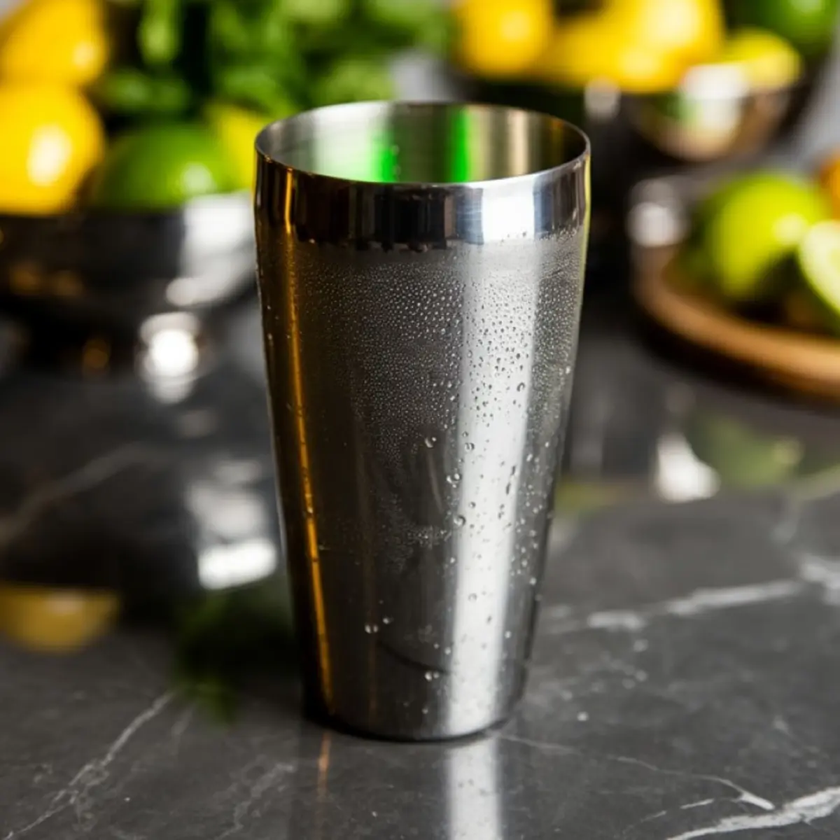 Metal cocktail shaker with condensation on a black marble counter, surrounded by blurred lemons, limes, and fresh herbs in the background. Halloween drink preparation scene.
