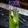 Green Halloween drink in a spider-themed tall glass with ice, rosemary sprig, and a cucumber spear, placed on a dark marble counter under bold “Green Witch Elixir” text.