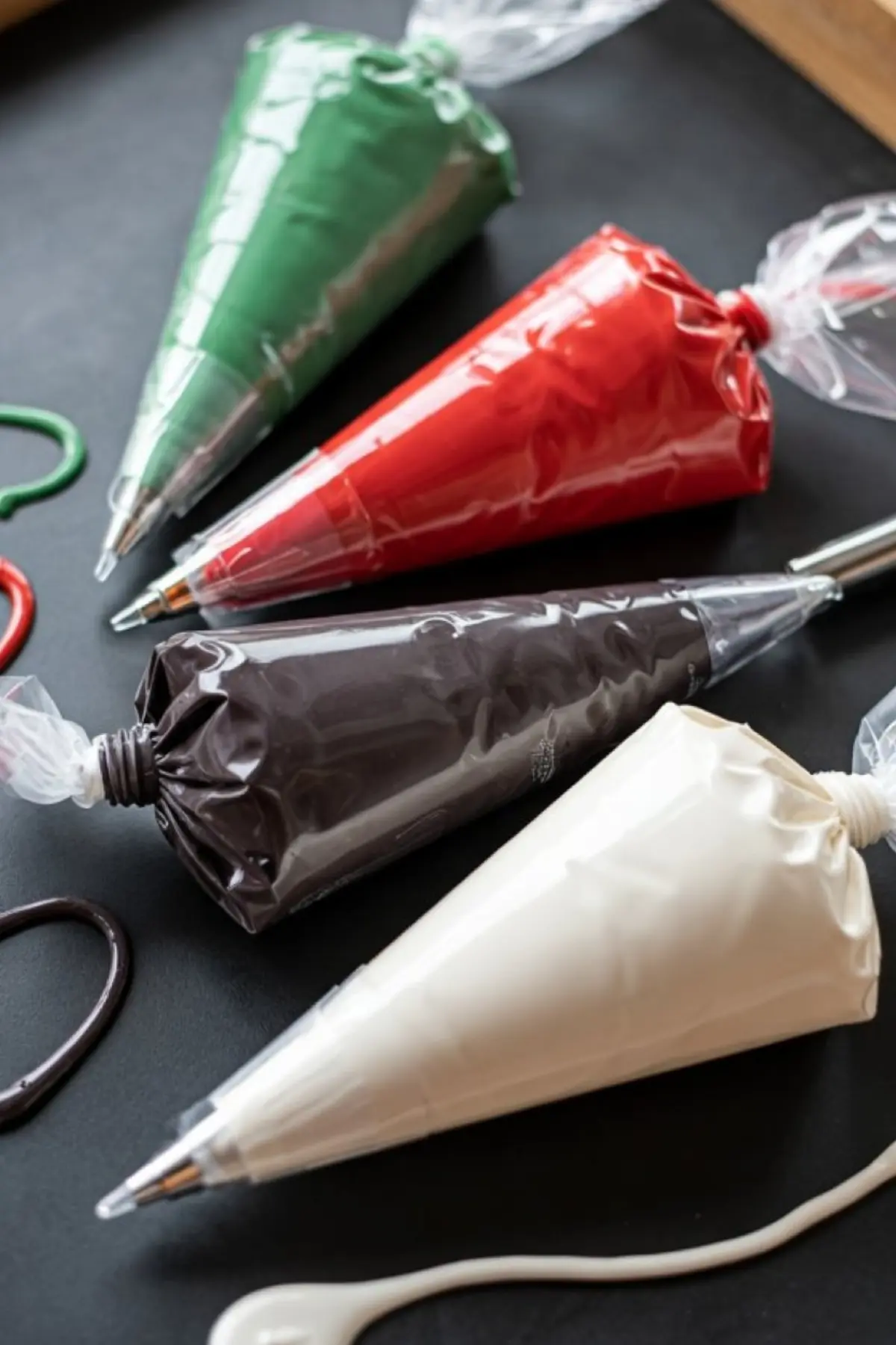 Four piping bags filled with red, green, white, and dark brown royal icing, arranged diagonally on a black surface for cookie decorating.