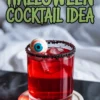 Spooky red Halloween cocktail in a glass with a black sugar rim, ice cubes, and a floating candy eyeball garnish, styled with pomegranate seeds on a coaster.