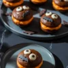 Chocolate whoopie pies with orange filling, decorated with candy eyes and bat cutouts, arranged on a black plate with bat-shaped confetti on a dark marble surface.