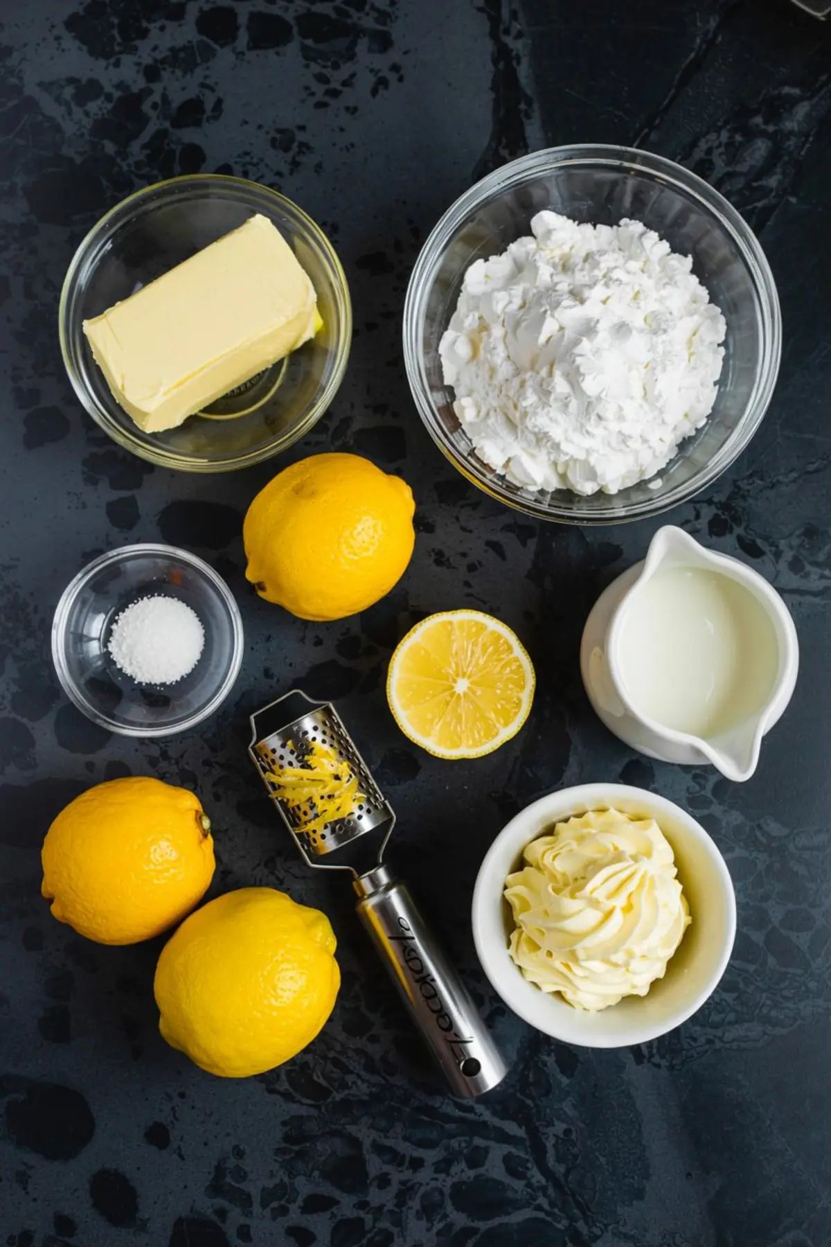 Flat lay of lemon buttercream frosting ingredients including fresh lemons, butter, powdered sugar, salt, heavy cream, lemon zest, and a lemon zester arranged on a dark surface.