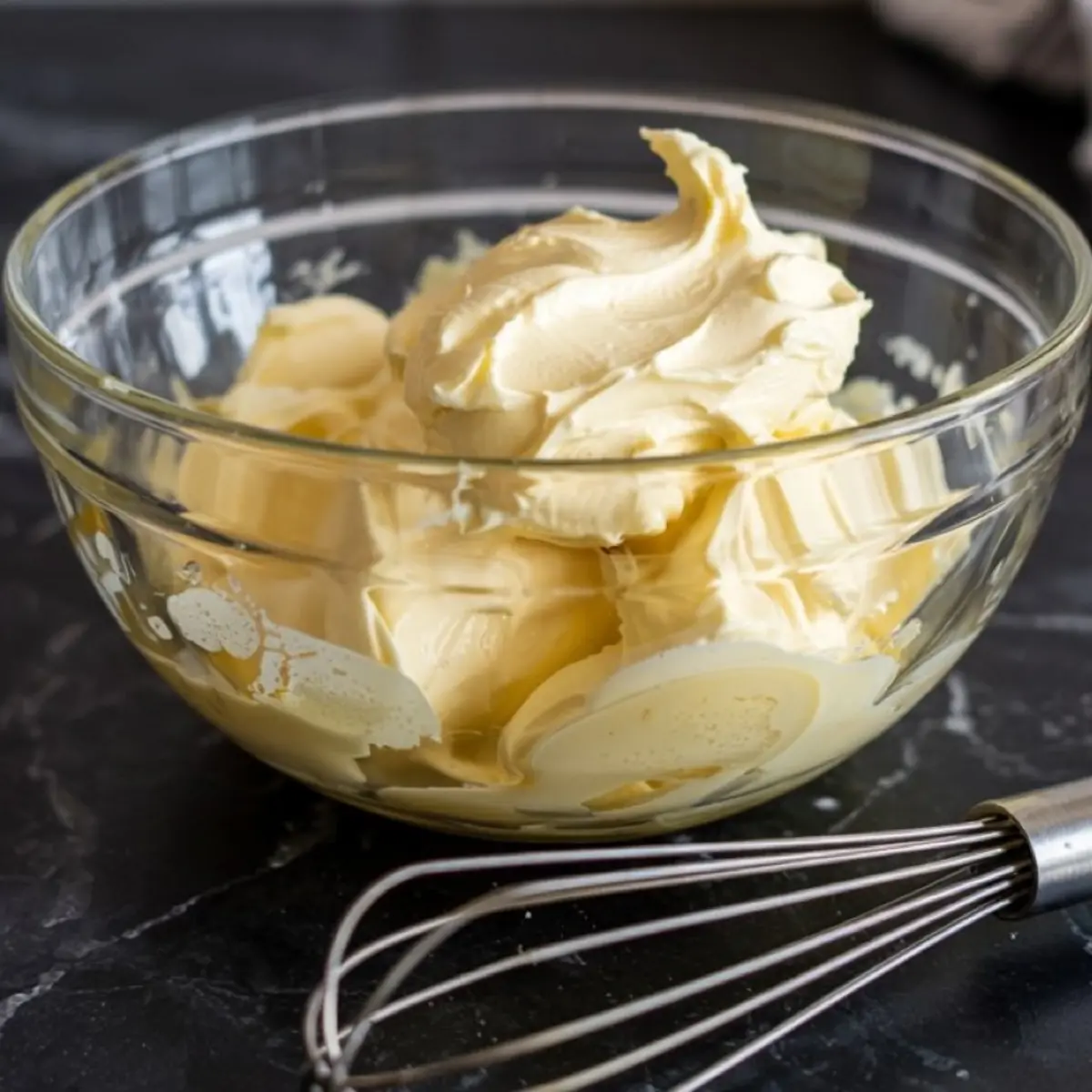 Thick and creamy lemon butter mixture whipped to soft peaks in a glass bowl, with a metal whisk placed on the black countertop.