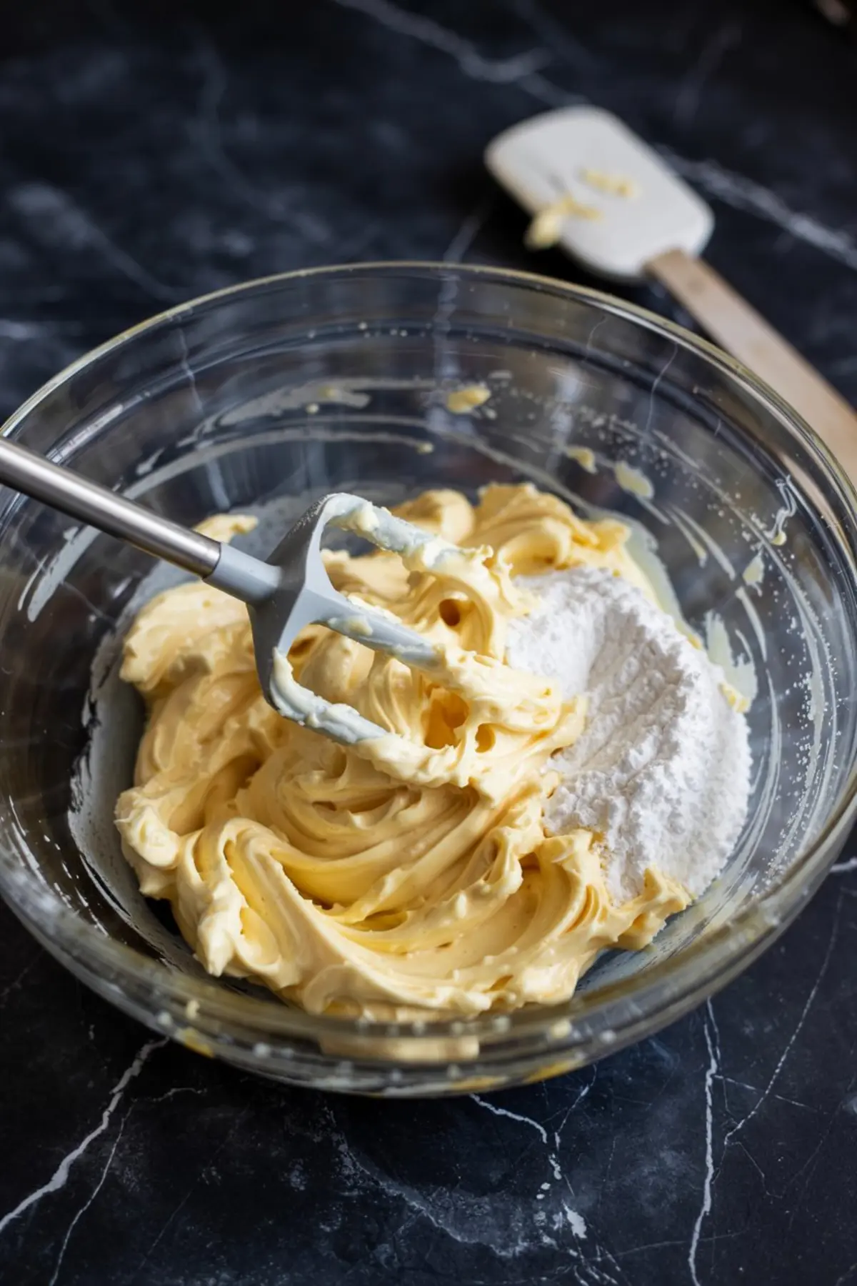 Glass bowl with partially mixed lemon buttercream frosting and powdered sugar, with a spatula resting nearby on a dark marble counter.