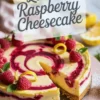 Lemon raspberry cheesecake with raspberry swirls and a slice removed, topped with fresh raspberries, lemon curls, and mint leaves, displayed on a pink plate with powdered sugar.