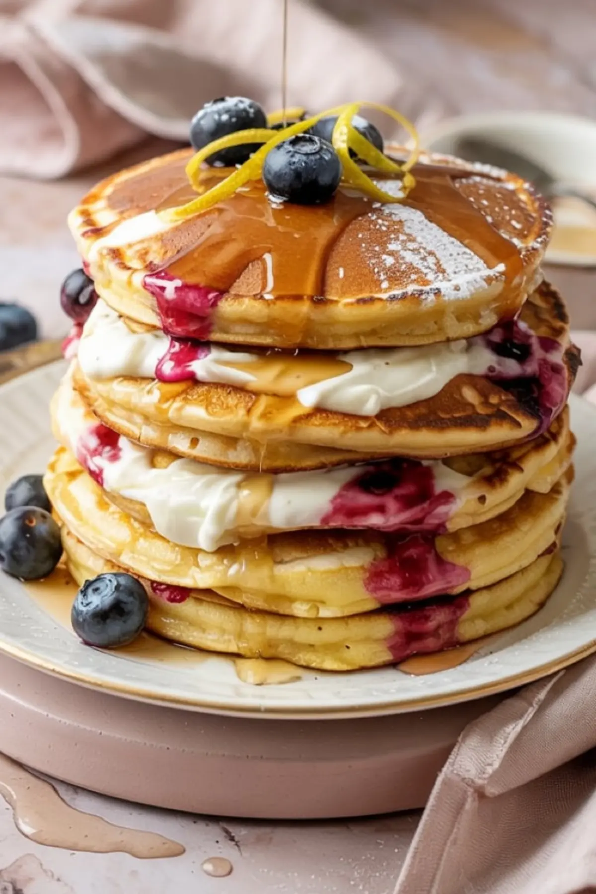 Tall stack of lemon ricotta pancakes layered with creamy filling and blueberry sauce, drizzled with syrup and garnished with fresh blueberries, lemon zest curls, and powdered sugar on a white plate.
