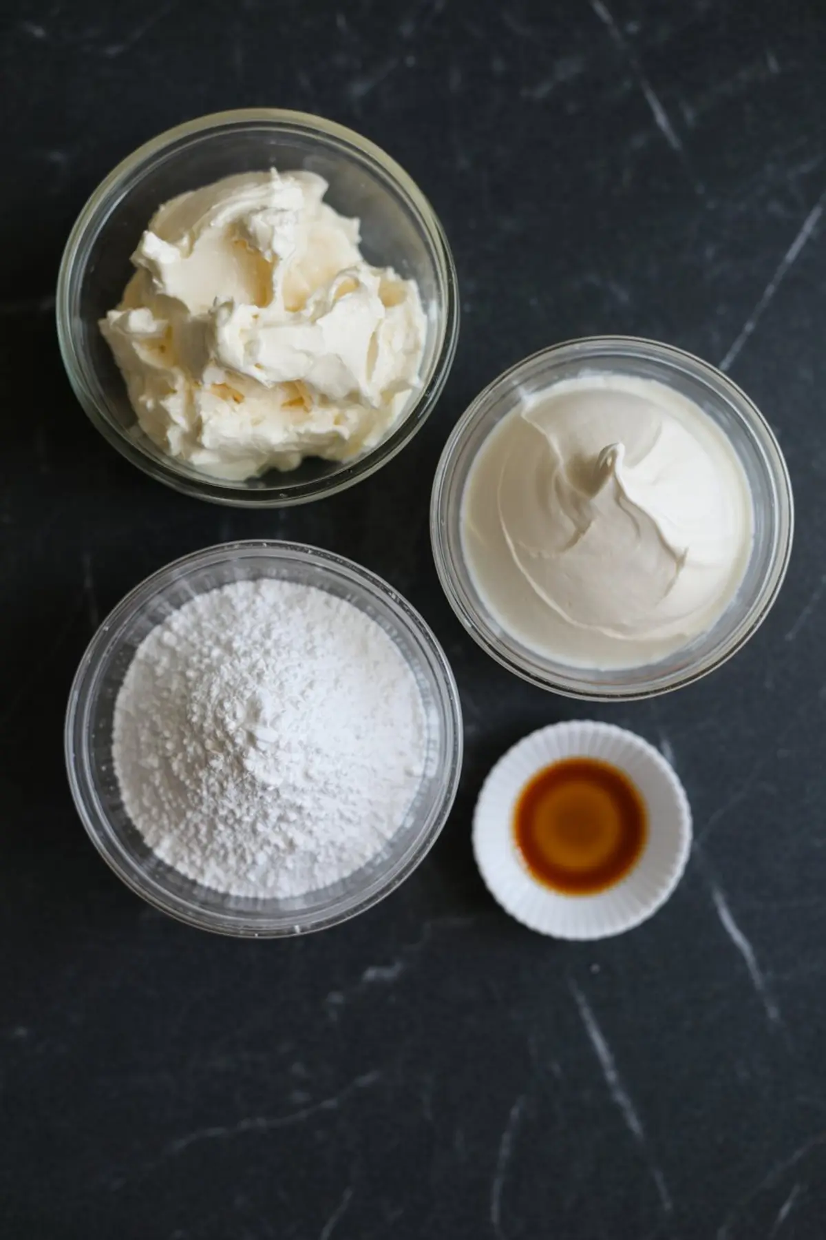 Overhead shot of mascarpone cheese, cream cheese, powdered sugar, and vanilla extract in clear bowls on a black marble countertop.
