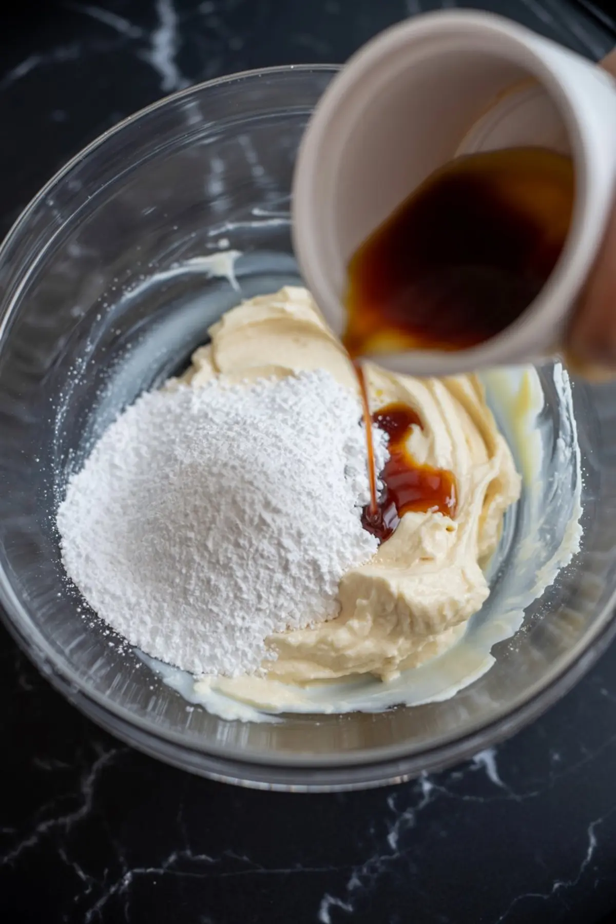 Close-up of vanilla extract being poured into a mixing bowl with powdered sugar and whipped cream cheese on a black counter.
