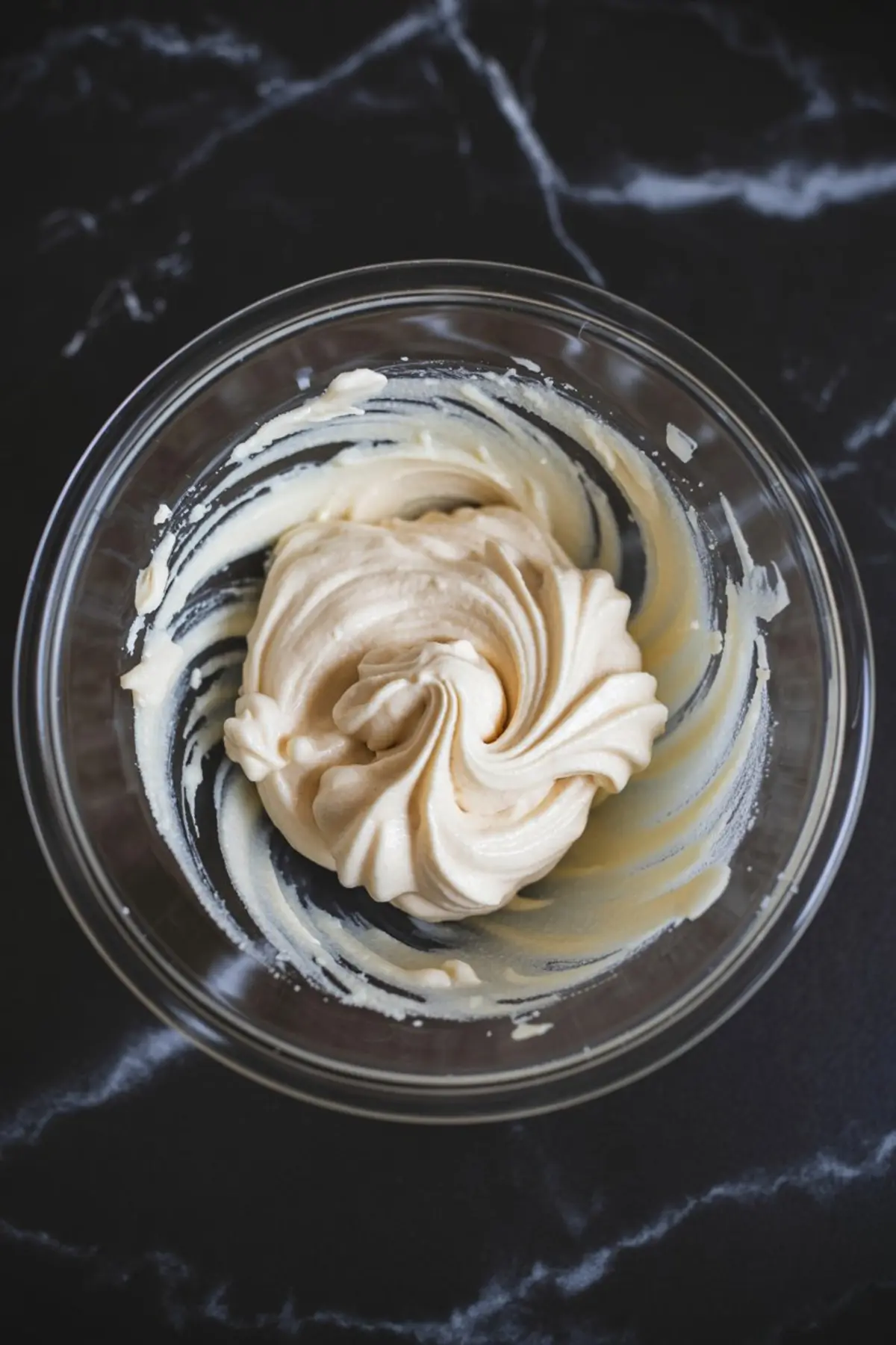 Fluffy mascarpone frosting swirled in a glass mixing bowl, with creamy texture and soft peaks against a dark marble background.
