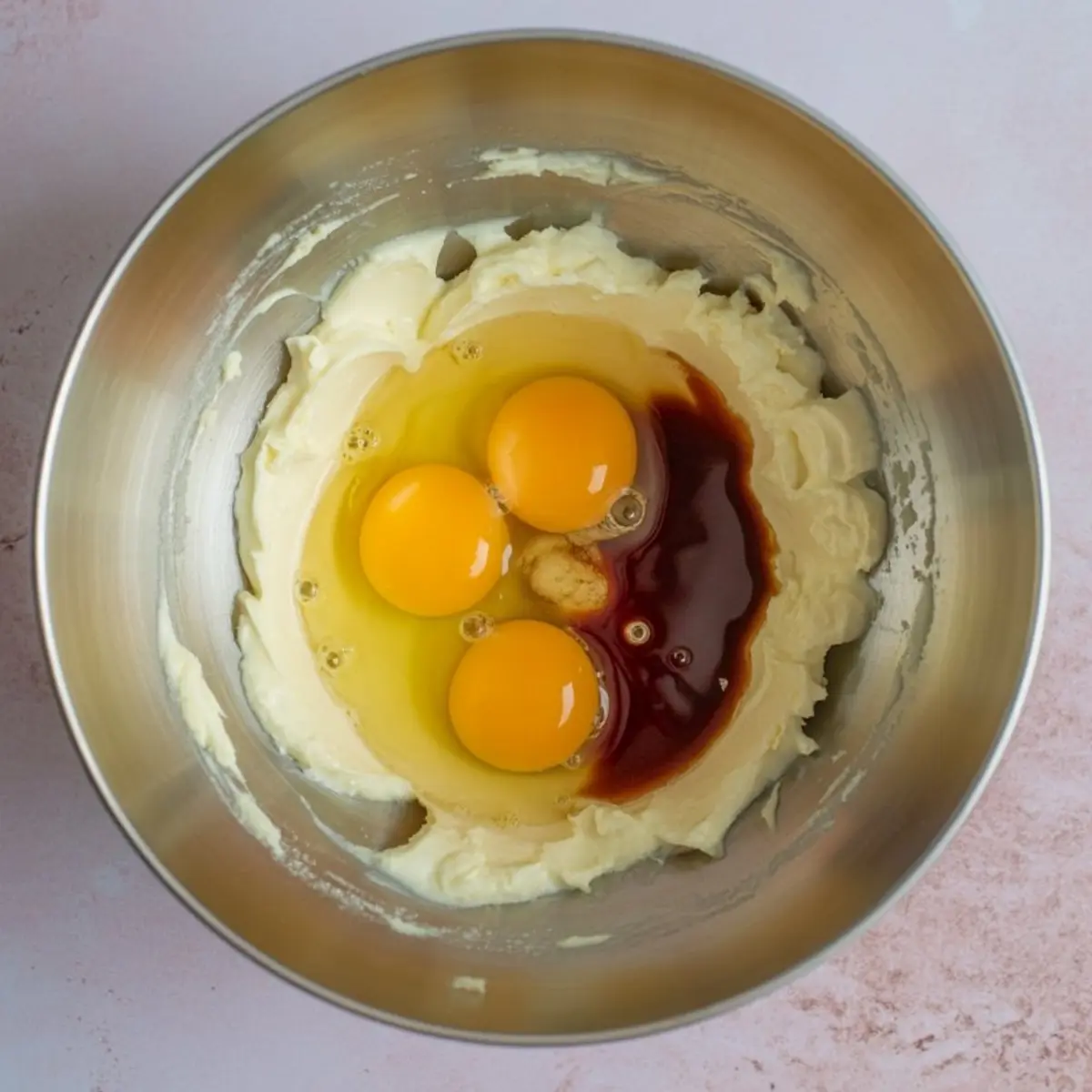 Metal mixing bowl with softened butter, three cracked eggs, vanilla extract, and sugar, prepared for cookie dough mixing on a pink background.
