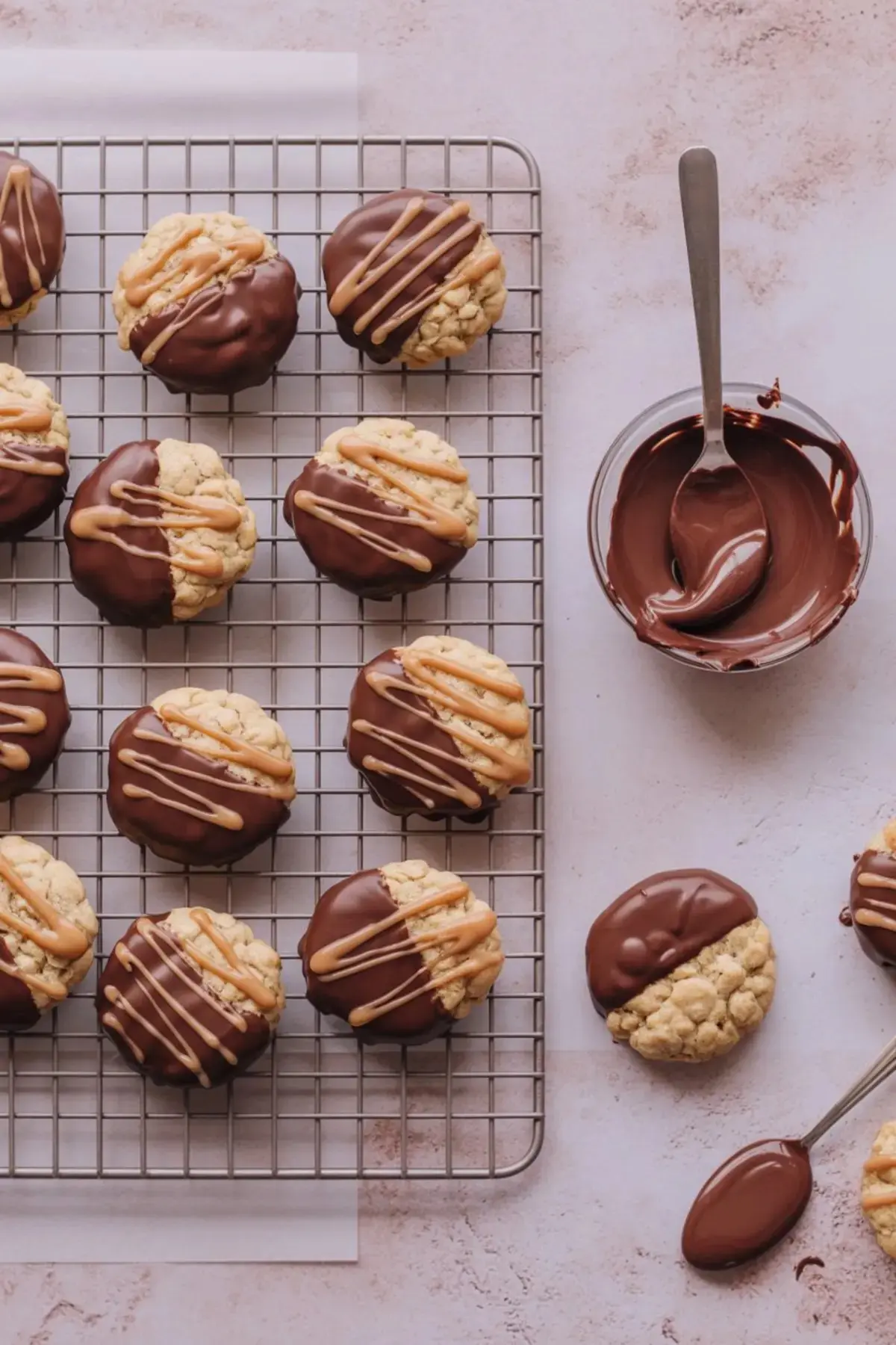 Cooling rack filled with oatmeal butterscotch cookies dipped in chocolate and drizzled with butterscotch, with a bowl of melted chocolate and a spoon beside it.
