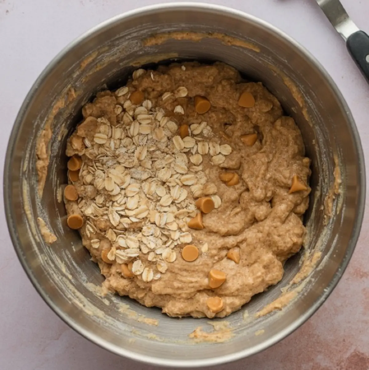 Cookie dough in a mixing bowl with rolled oats and butterscotch chips added on top, ready to be mixed for baking.
