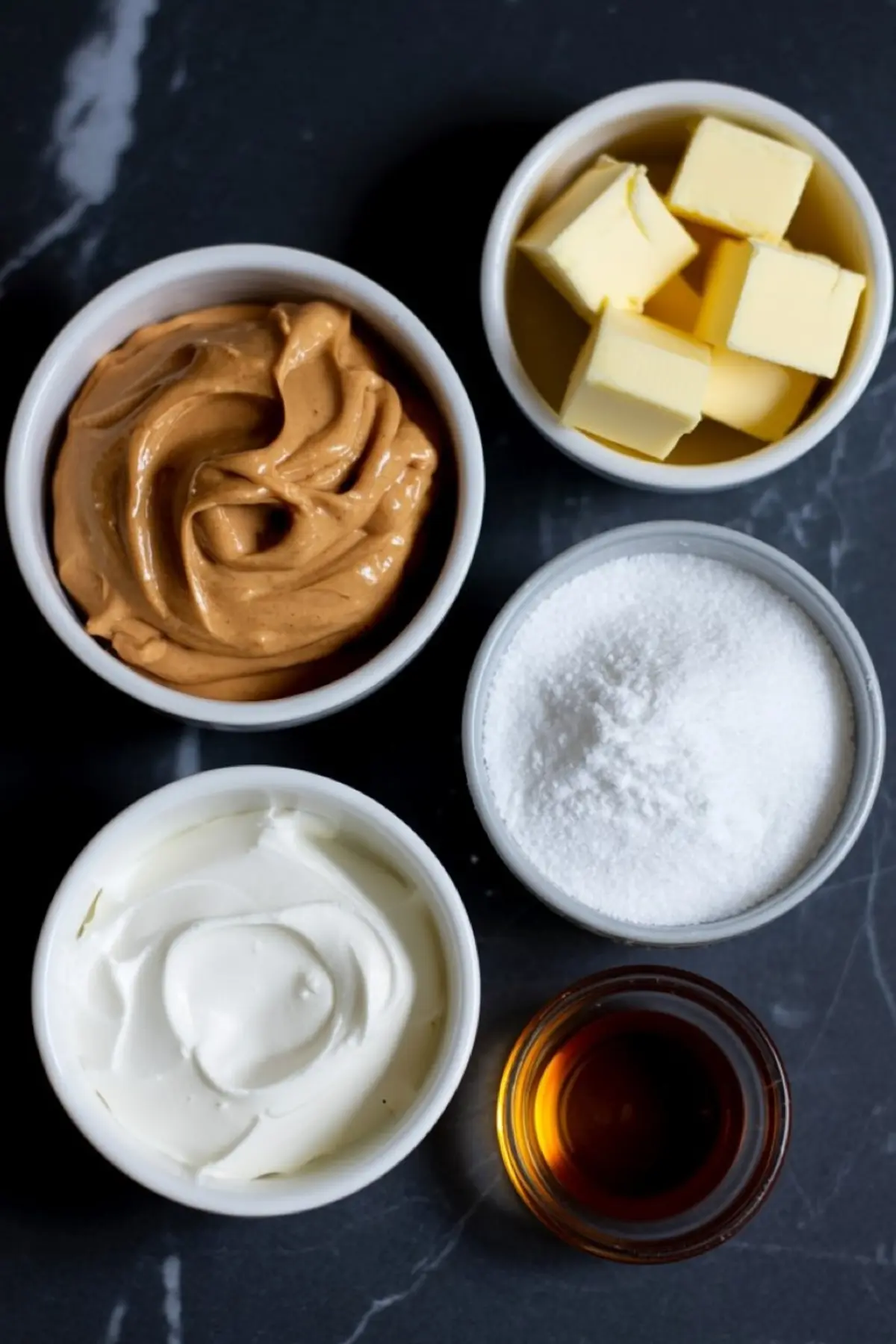 Top-down view of five small bowls on a dark marble surface holding peanut butter, cubed butter, powdered sugar, sour cream, and vanilla extract as ingredients for peanut butter frosting.
