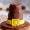 Close-up of a chocolate-covered pilgrim hat cookie with a yellow fondant band and buckle set on a textured light pink surface.