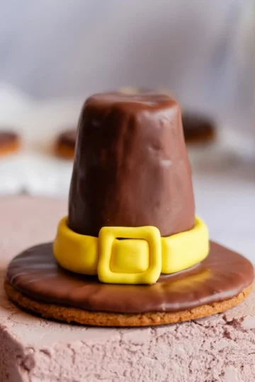 Close-up of a chocolate-covered pilgrim hat cookie with a yellow fondant band and buckle set on a textured light pink surface.