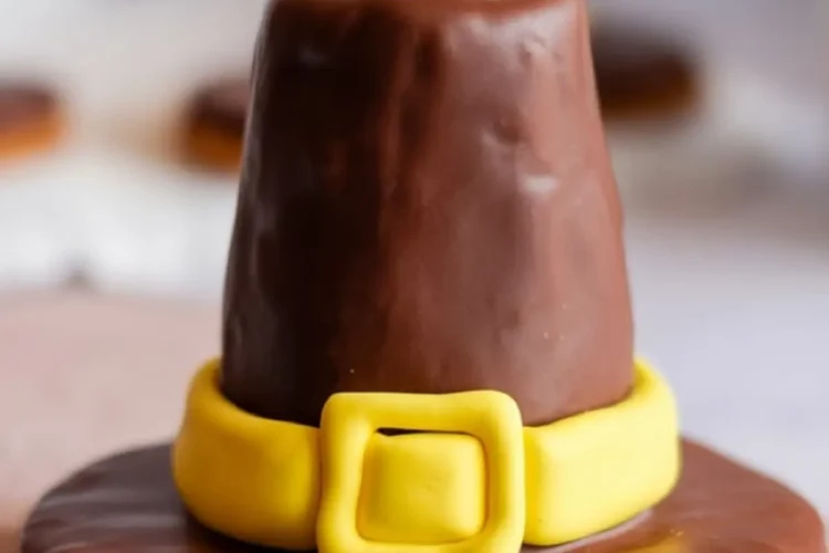 Close-up of a chocolate-covered pilgrim hat cookie with a yellow fondant band and buckle set on a textured light pink surface.