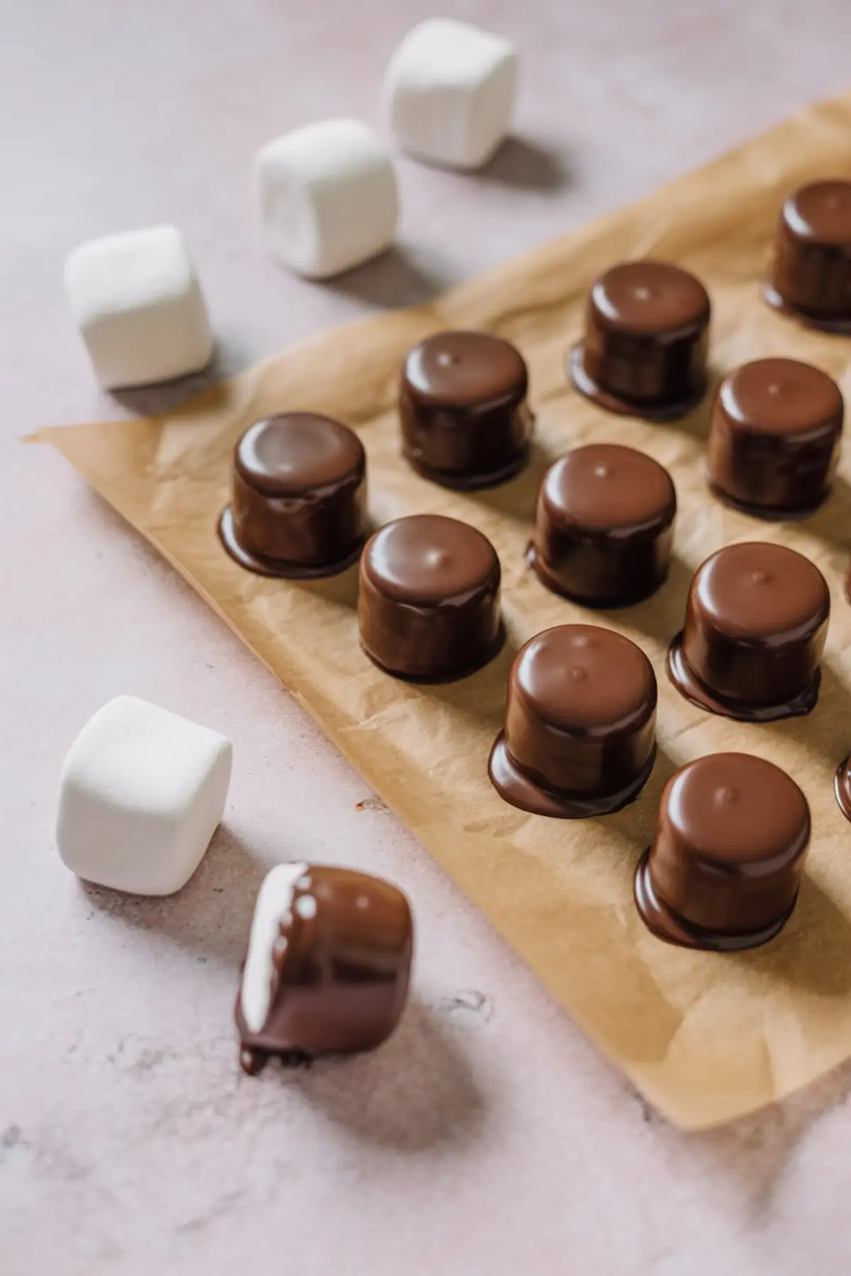 Partially dipped marshmallows coated in chocolate set on a piece of parchment paper with plain marshmallows nearby, arranged in neat rows for cooling.
